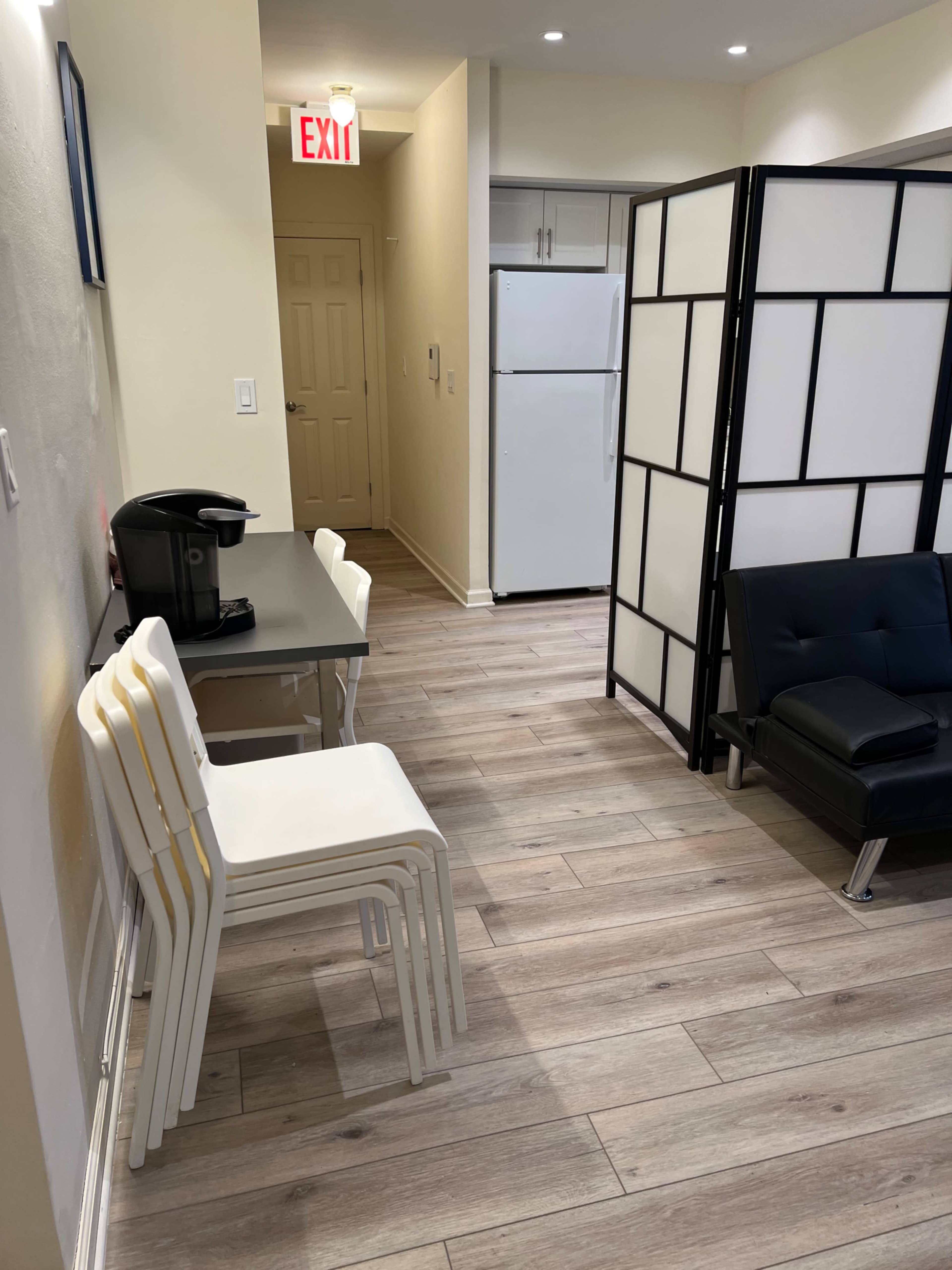 A narrow hallway with a table and stacked white chairs on one side and a refrigerator and partitioned area on the other.
