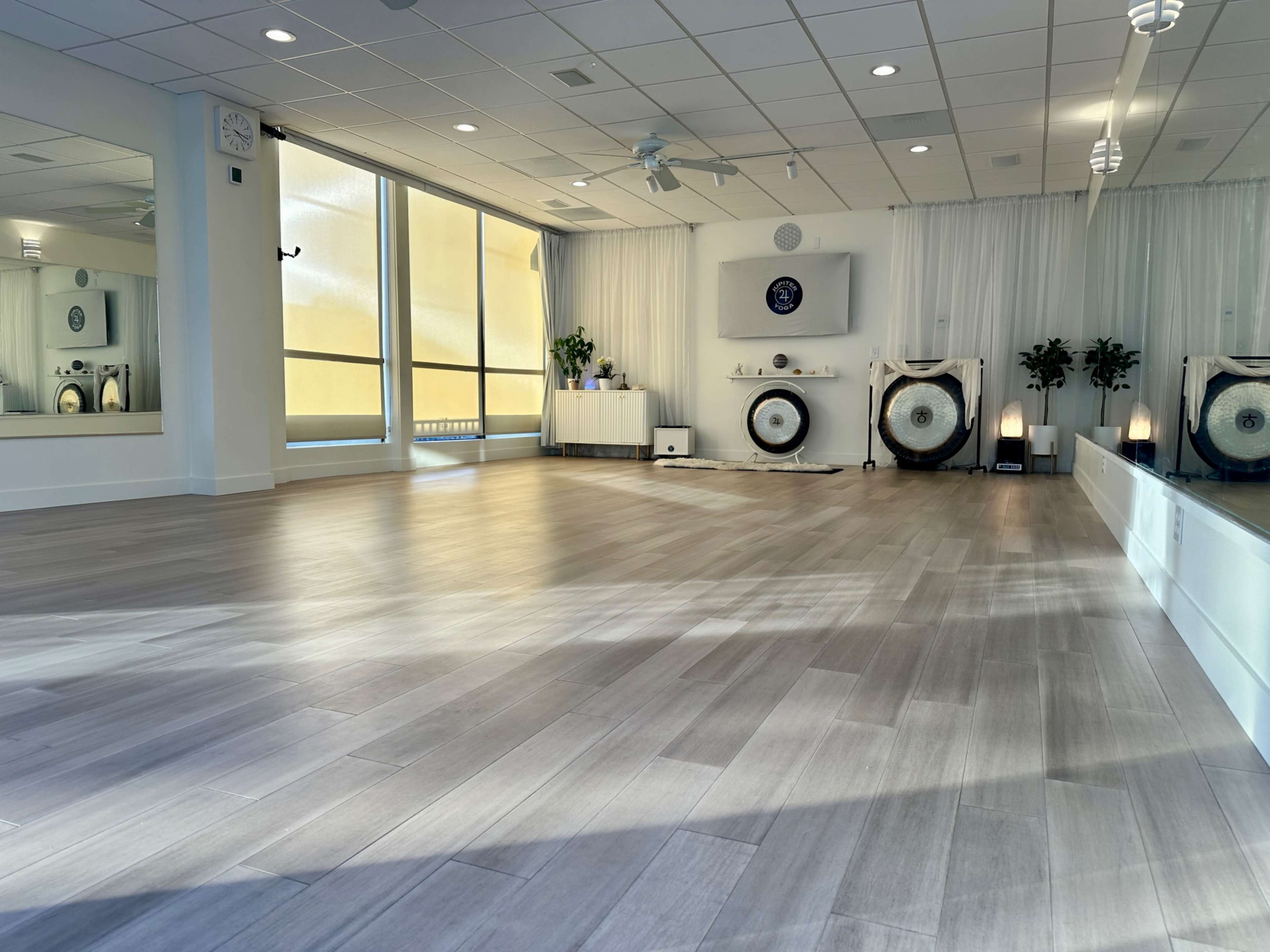 A spacious room with wooden flooring, large windows providing natural light, and a minimalistic setup featuring gongs and plants along with white drapes and furniture.
