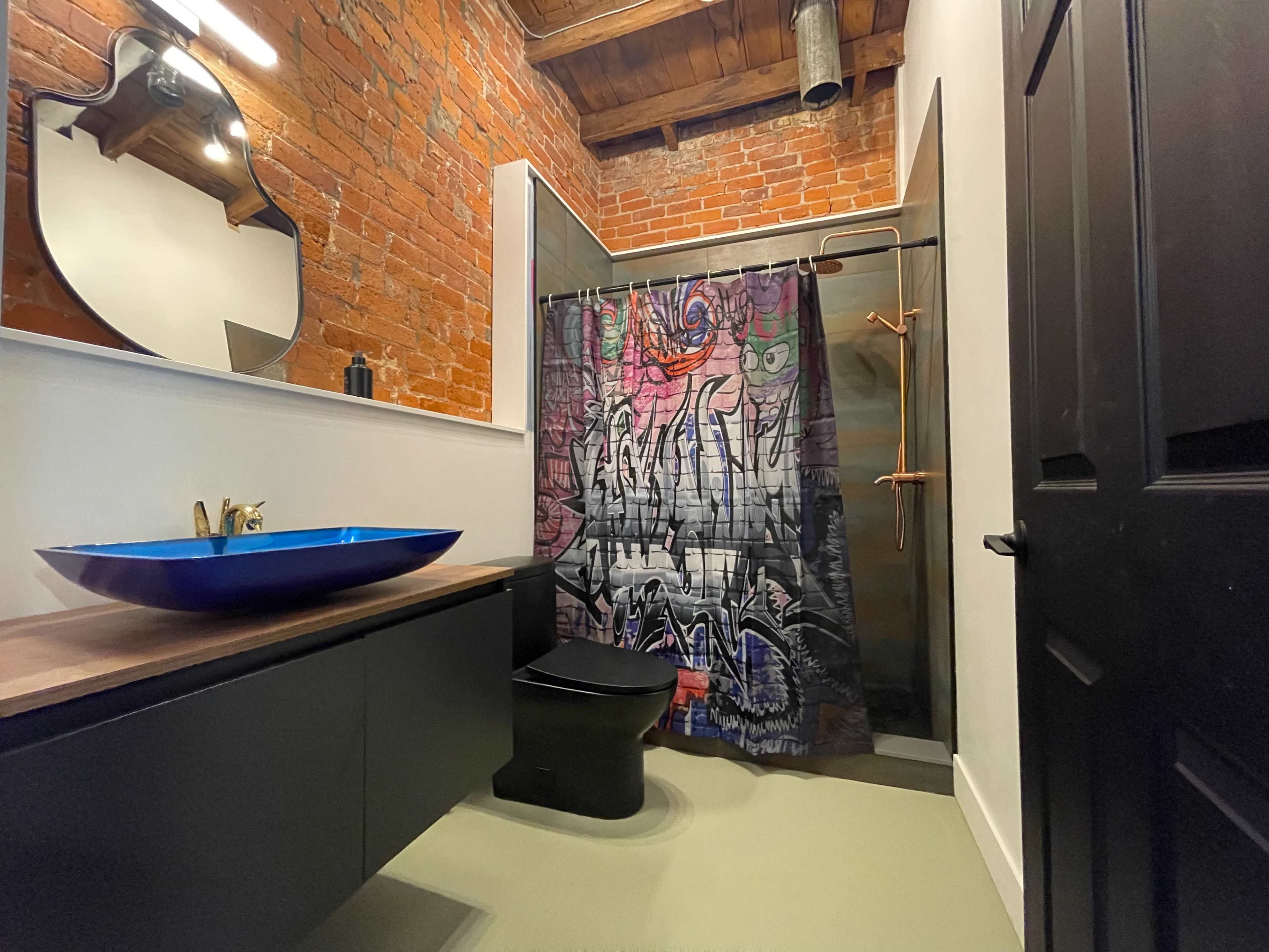 A bathroom with exposed brick walls, a modern black toilet, a large blue sink, and a colorful graffiti-style shower curtain.
