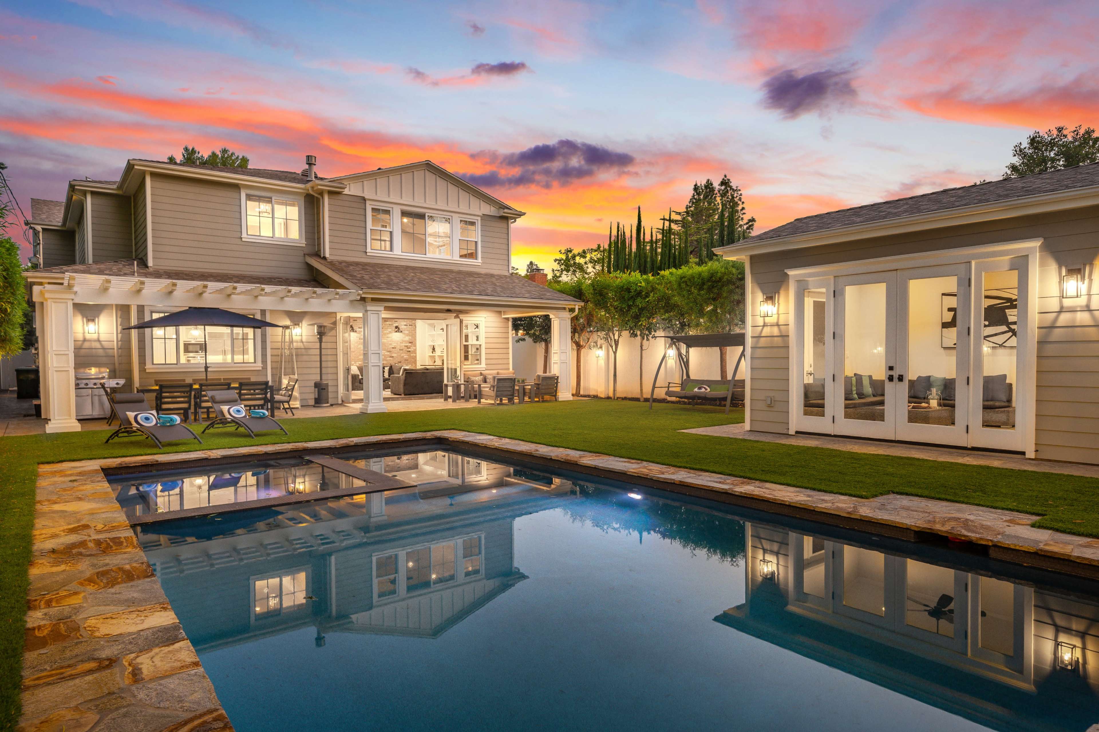 A modern two-story house with a landscaped backyard featuring a swimming pool and a colorful sunset sky.