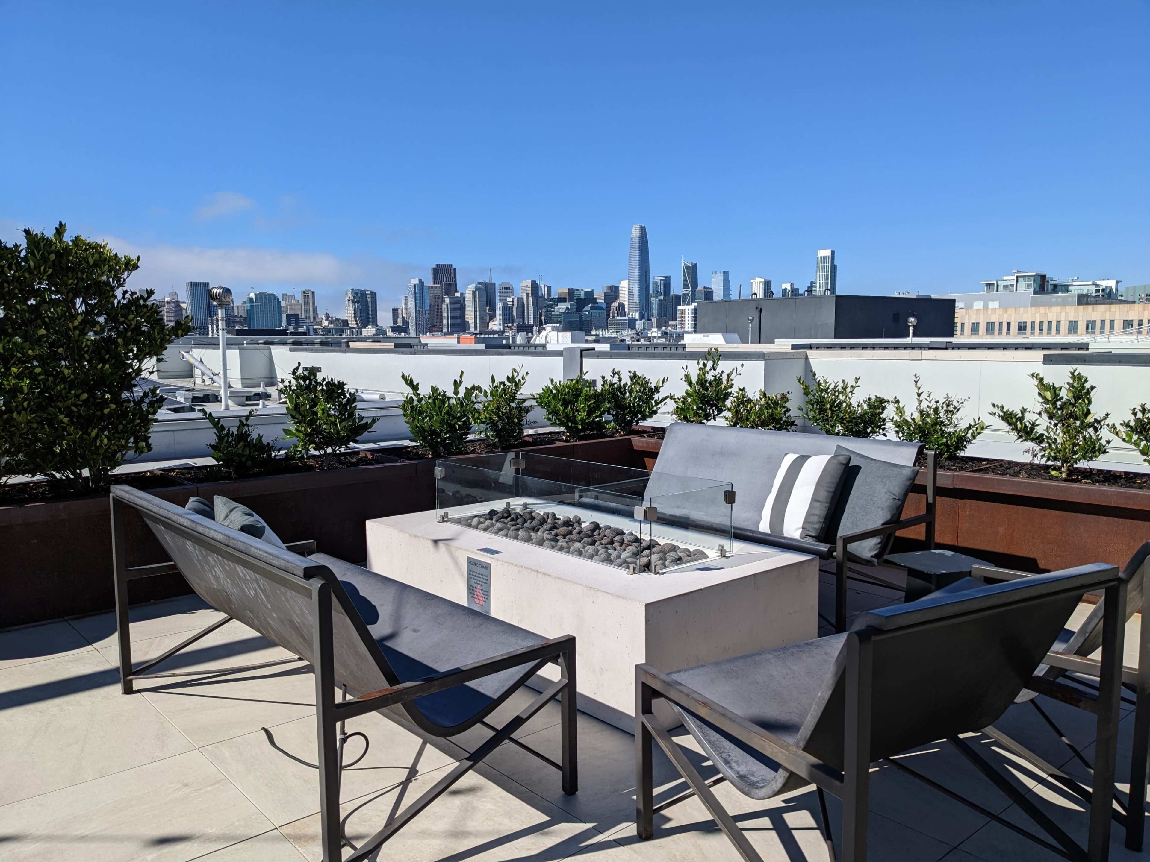 A rooftop patio features a fire pit surrounded by chairs and a view of a city skyline.