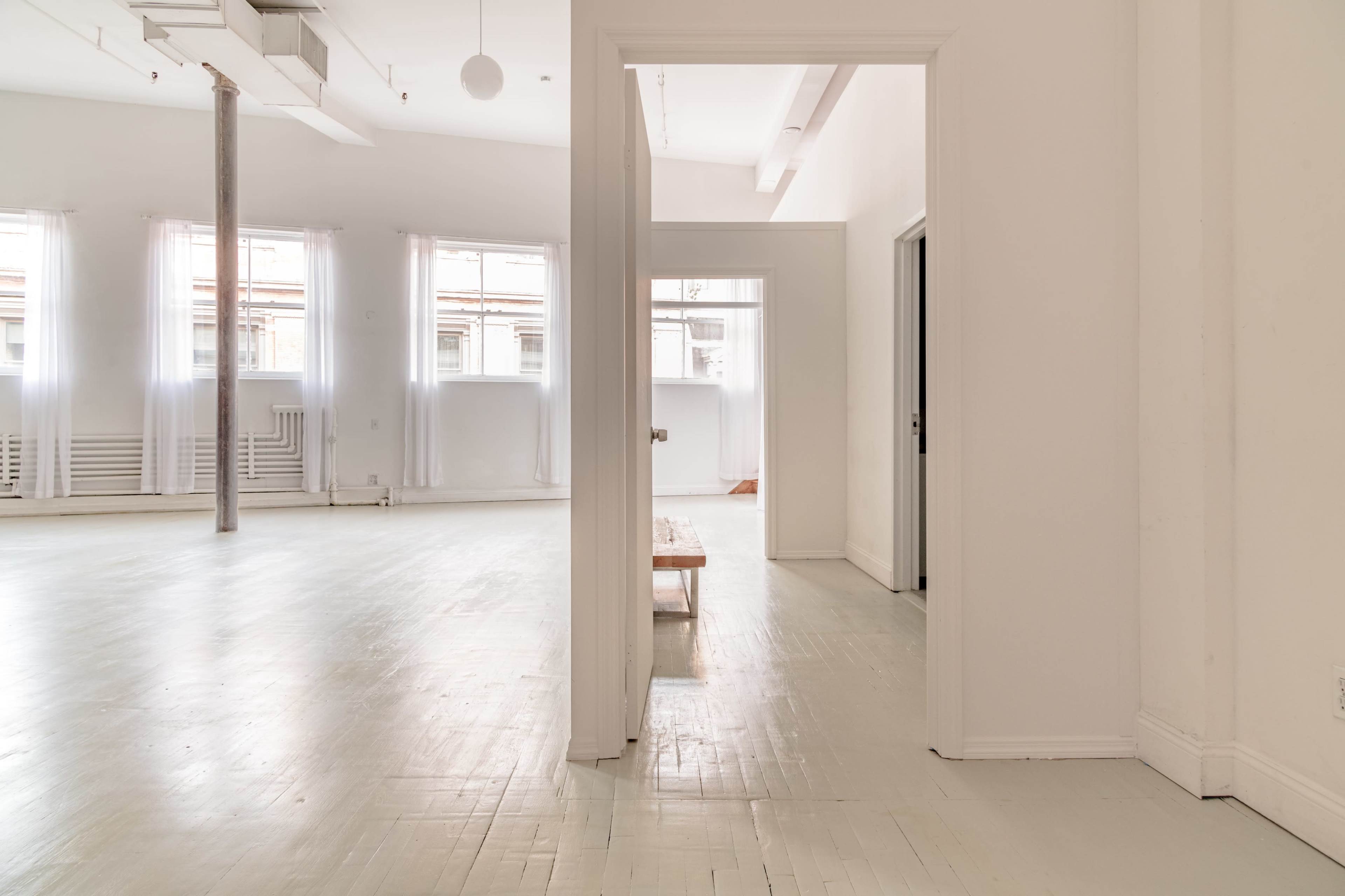 A light-filled, minimalist interior features white walls, a wooden bench, and multiple doorways leading to adjoining rooms.