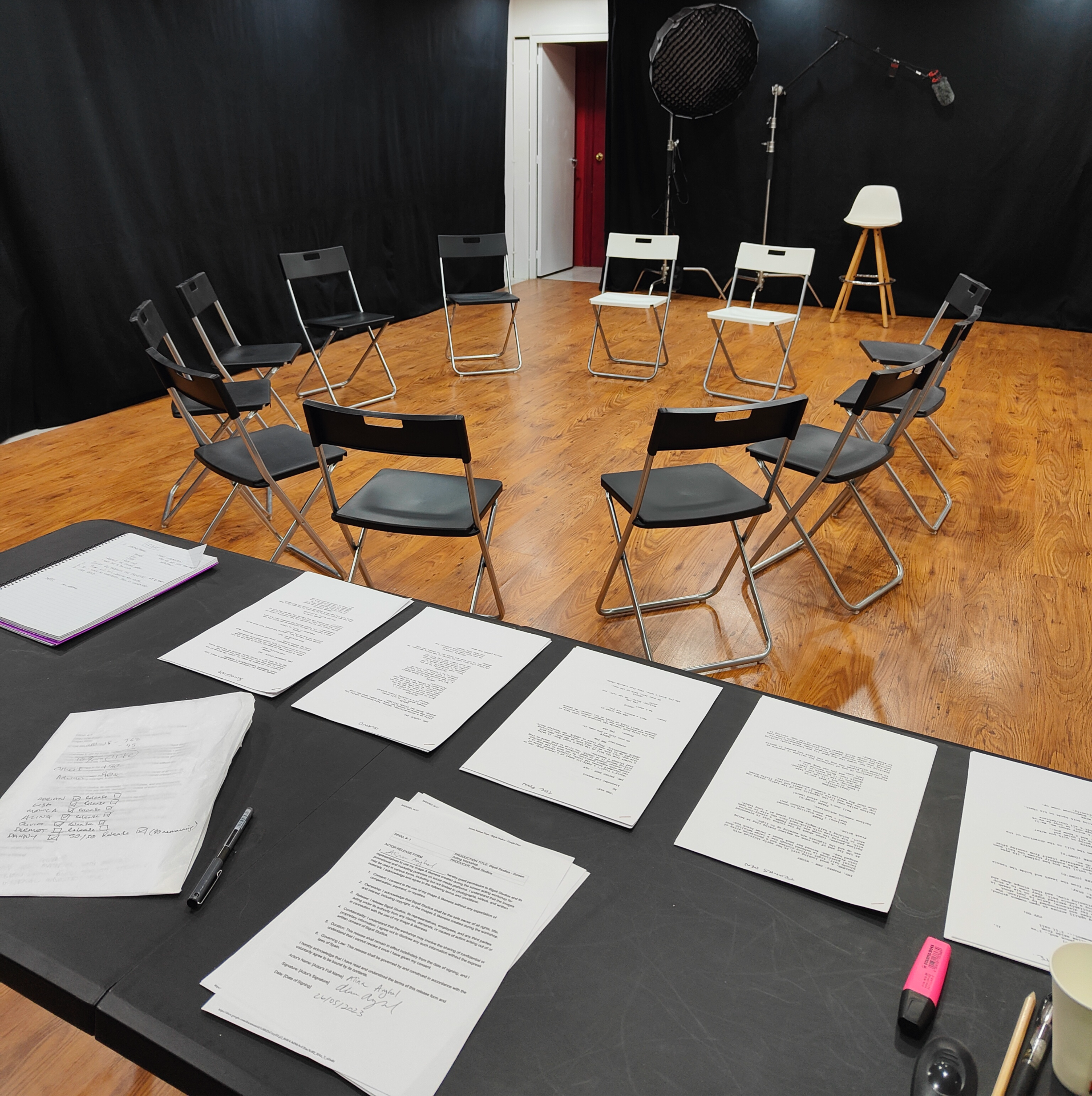 The image shows a rehearsal space with a circular arrangement of black chairs on a wooden floor, a table covered with scripts and paperwork, and a microphone setup in the background.
