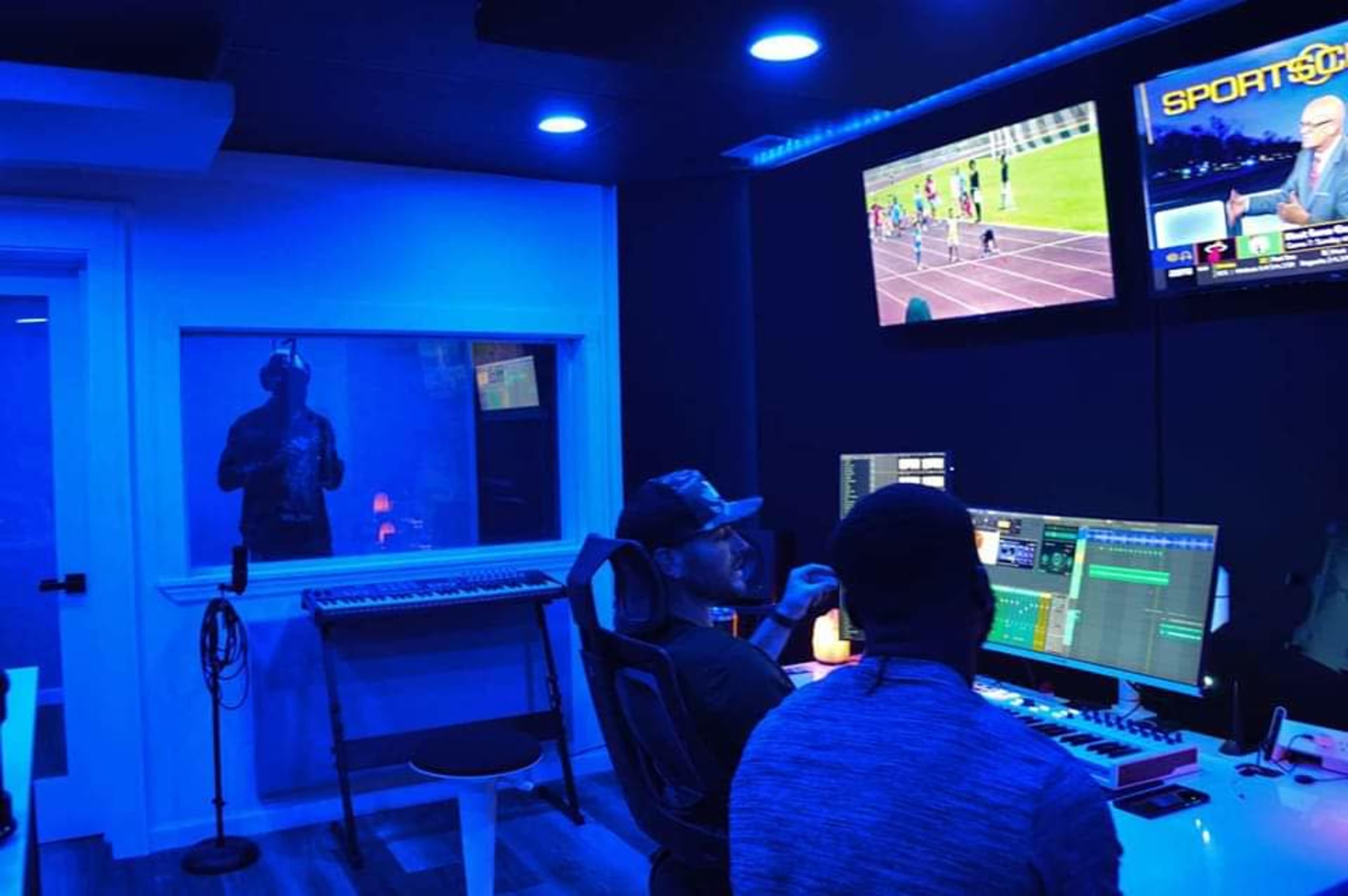 A music studio with two individuals working at a computer, while a third person is visible in a soundproof booth, and a television displays a sports event in the background.