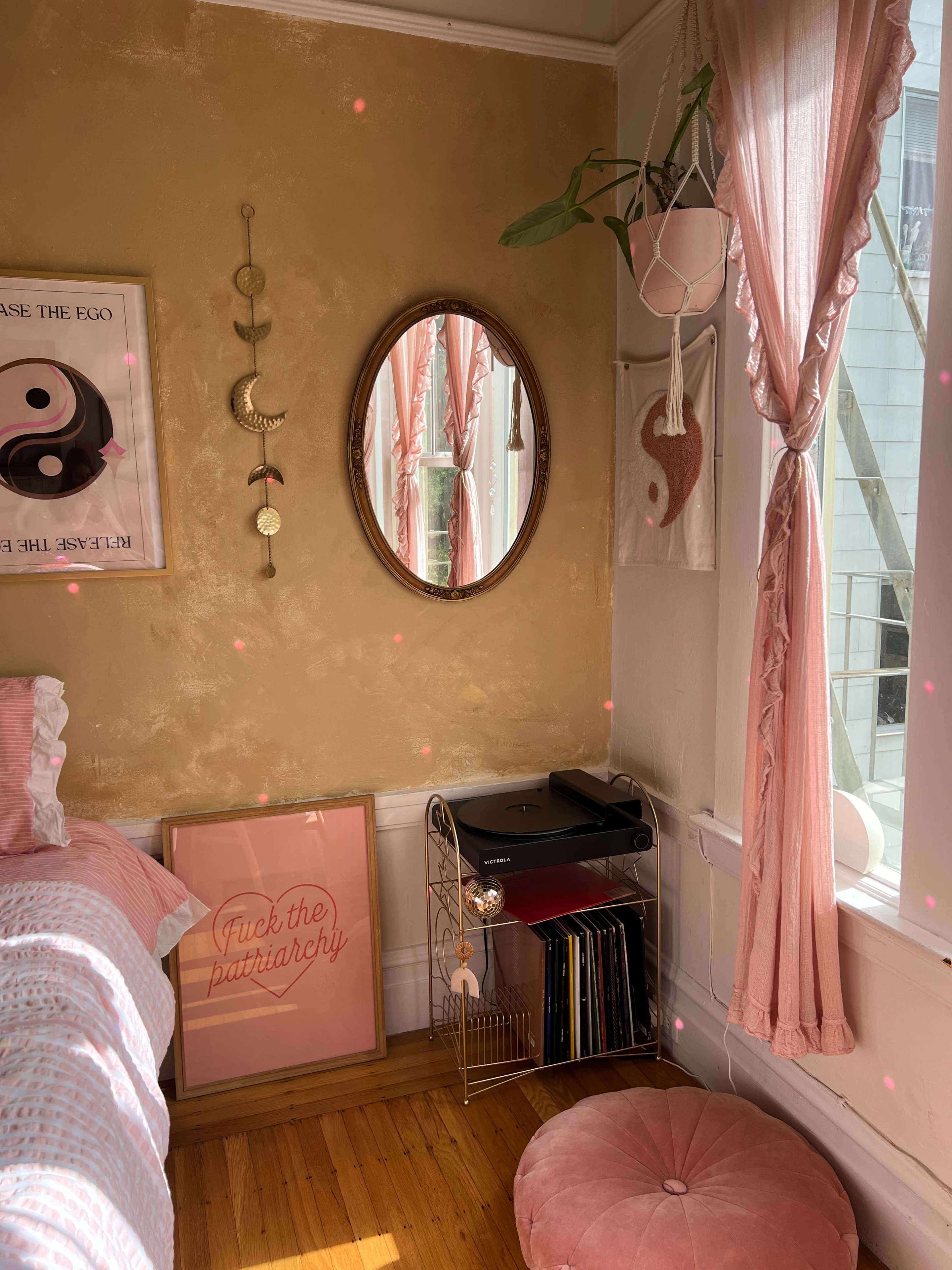A cozy bedroom corner featuring a bed with pink bedding, a round mirror, a decorative wall hanging, and a shelf with a printer and colorful files.