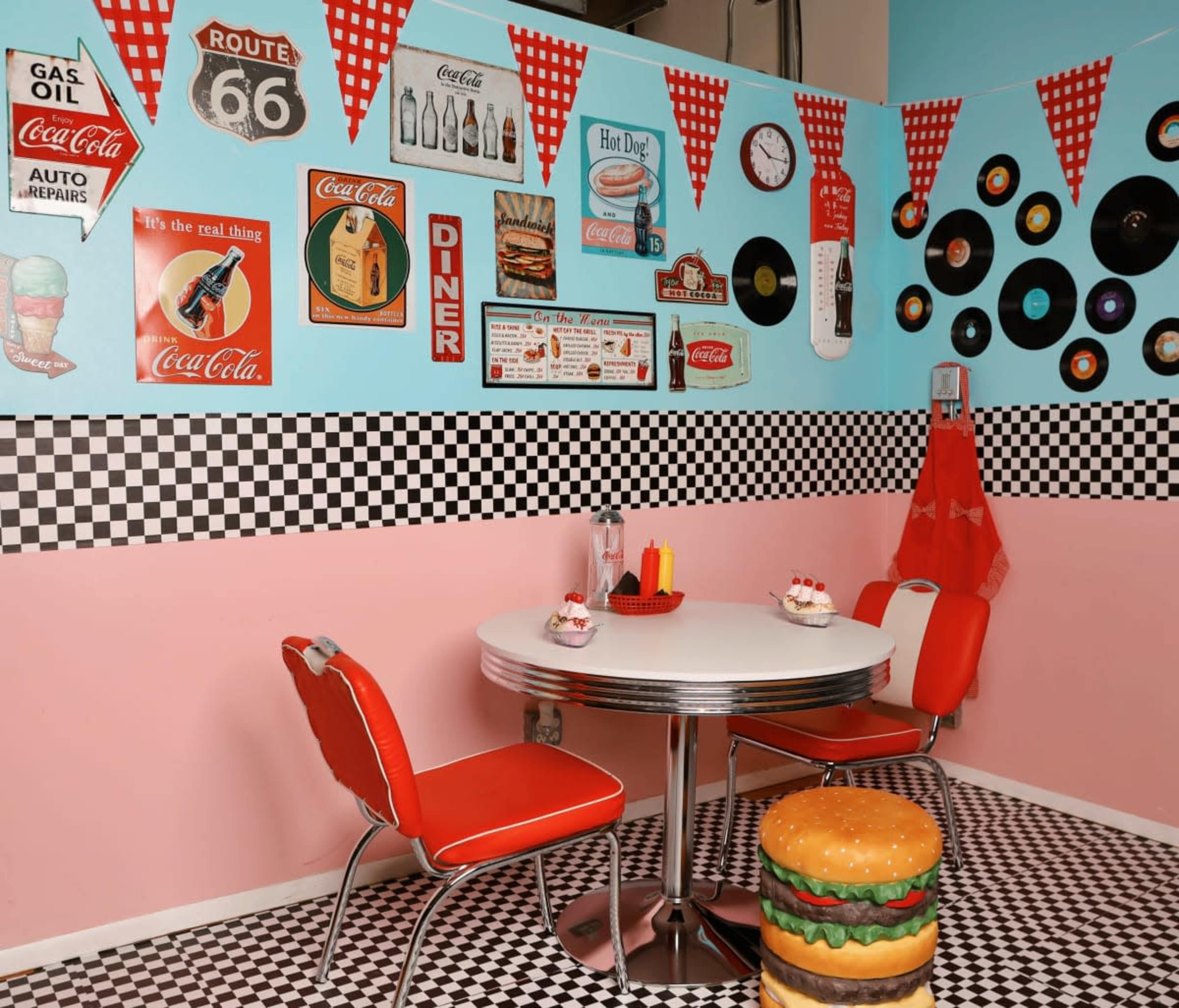 The image shows a retro diner-themed room with checkered flooring, a round table, red chairs, and various vintage Coca-Cola and Route 66 decorations on the walls.