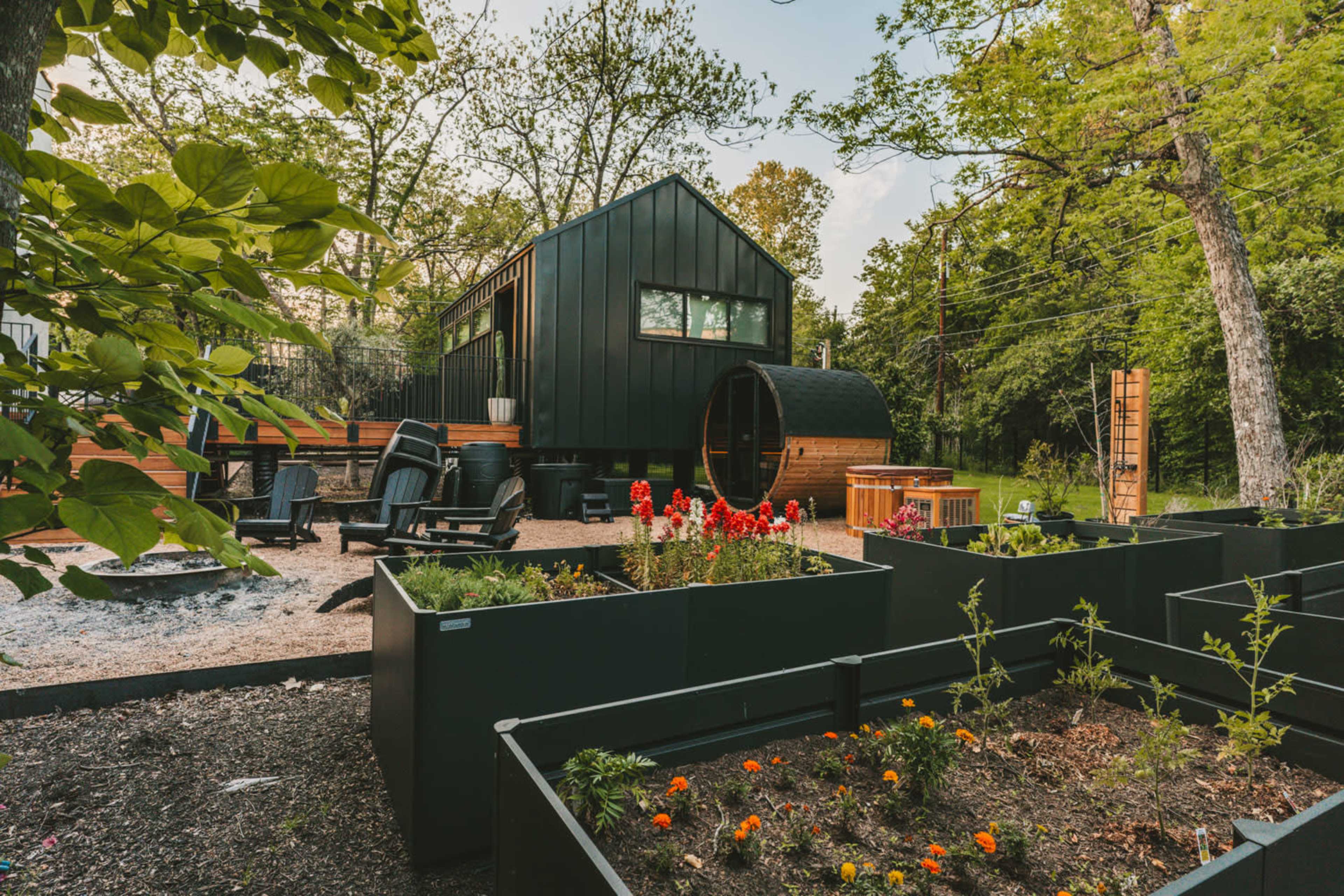 The image showcases a modern black tiny house surrounded by garden beds, a fire pit, and outdoor seating in a wooded area.