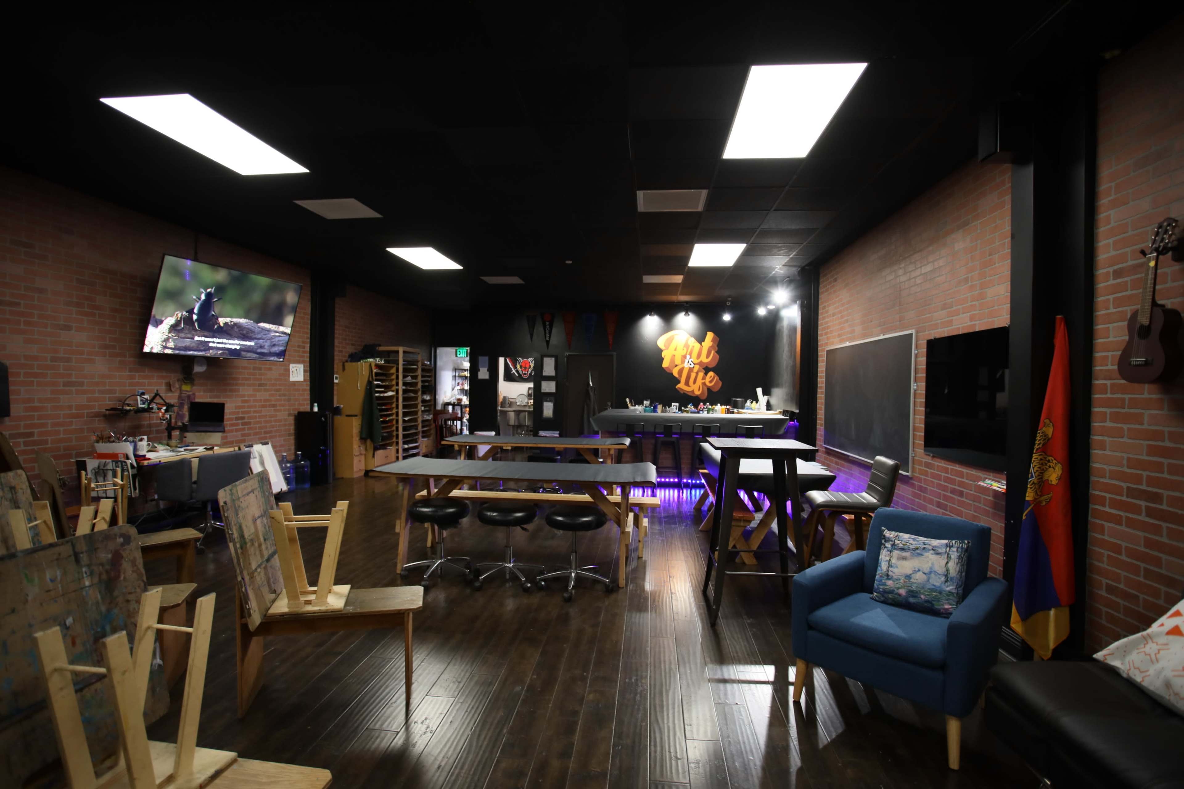 The image shows an art studio with wooden floors, exposed brick walls, and various tables set up for creative work, featuring a bar area illuminated with colorful lights.