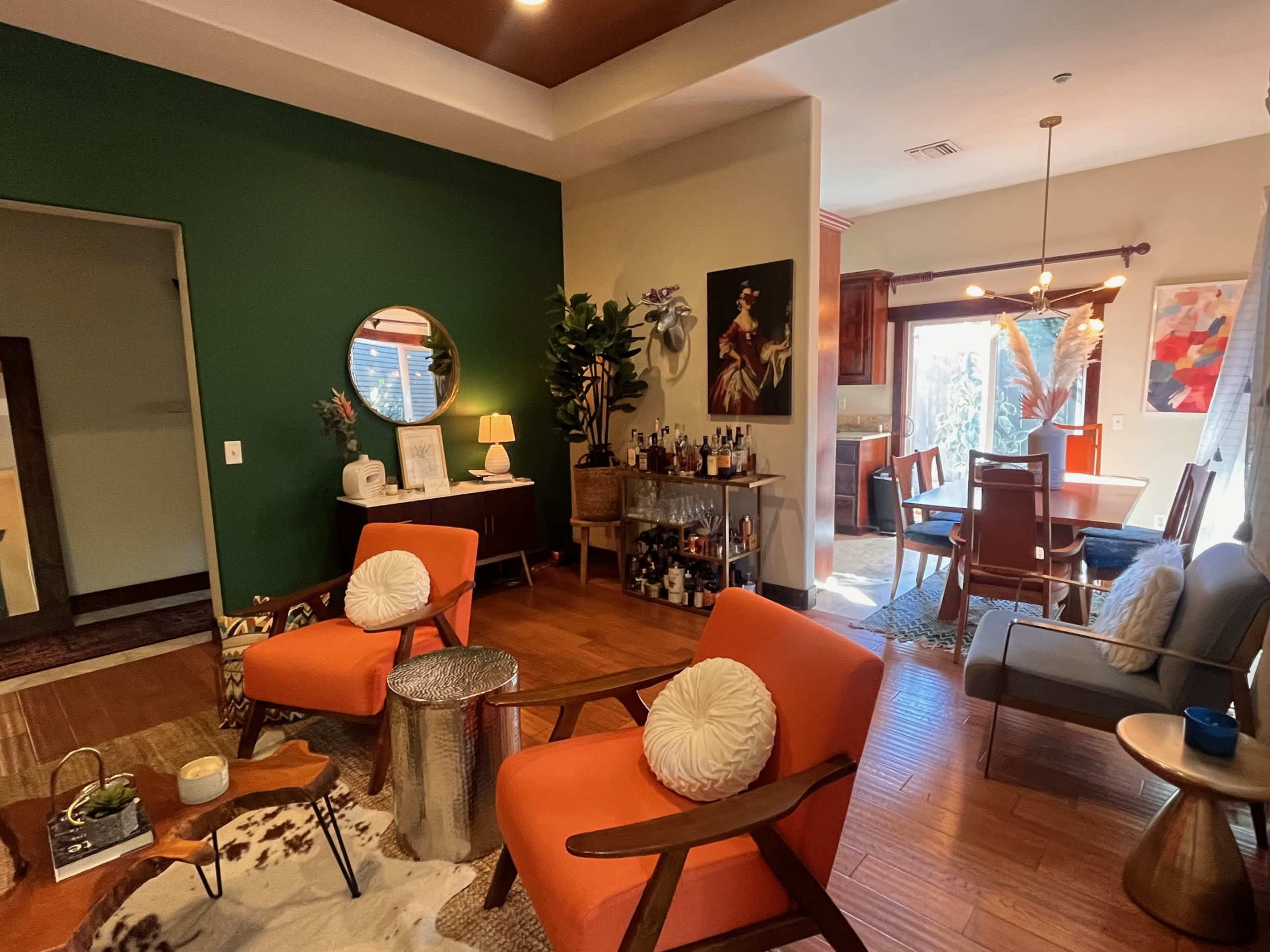The image shows a modern living area featuring orange accent chairs, a round mirror, a bar station, and a dining area with large windows.