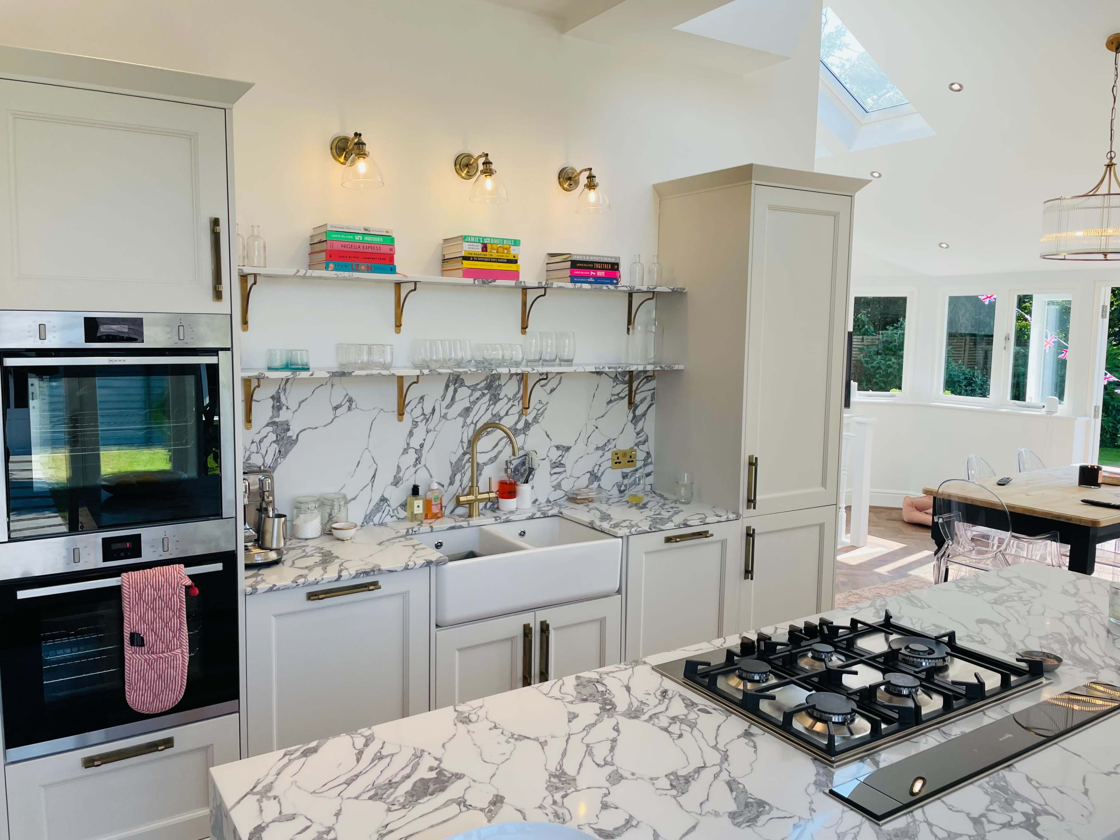A modern kitchen featuring marble countertops, a farmhouse sink, wall-mounted shelves with colorful books, and a gas stove.
