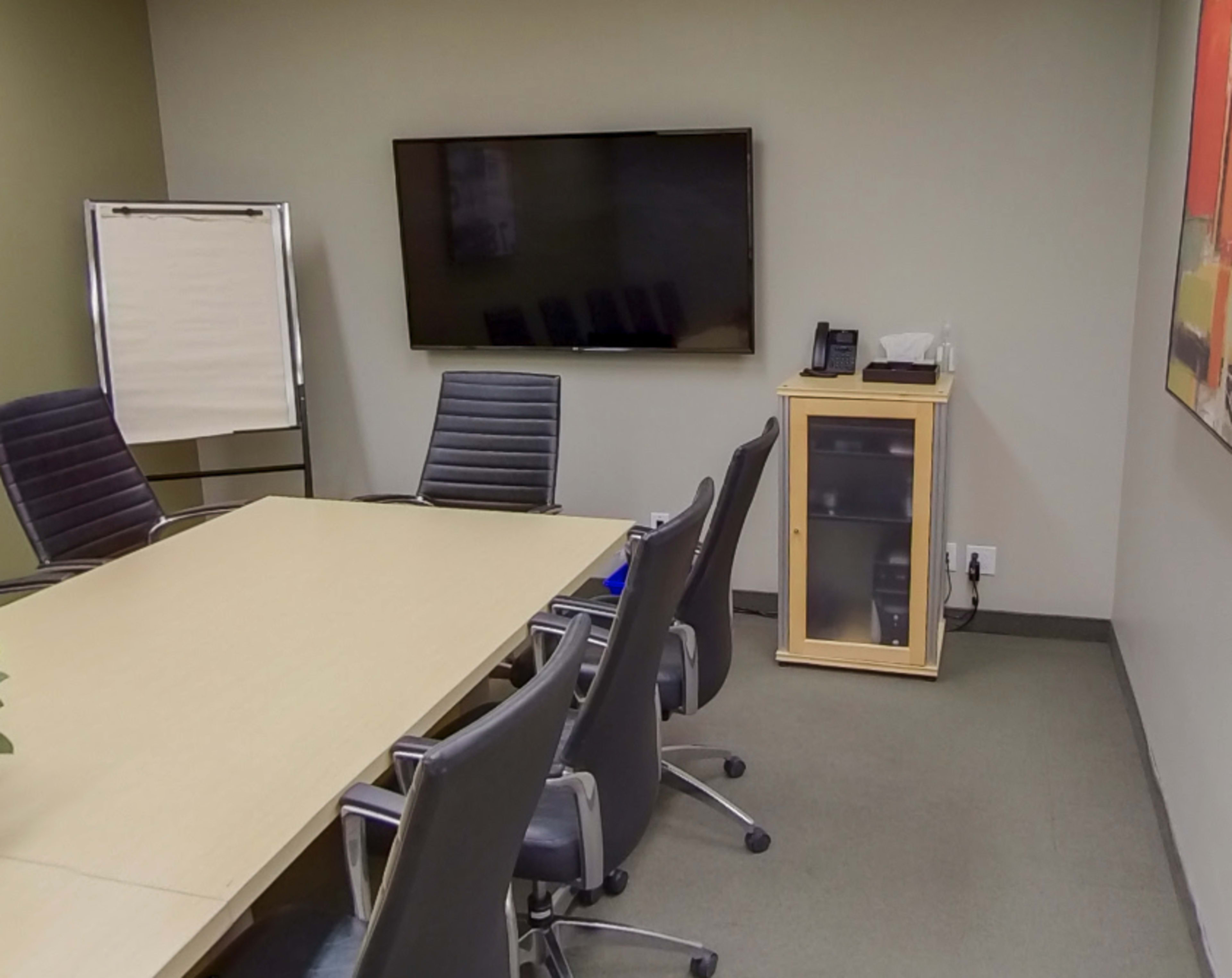 A conference room features a long table, several rolling chairs, a large television mounted on the wall, and a small cabinet in the corner.
