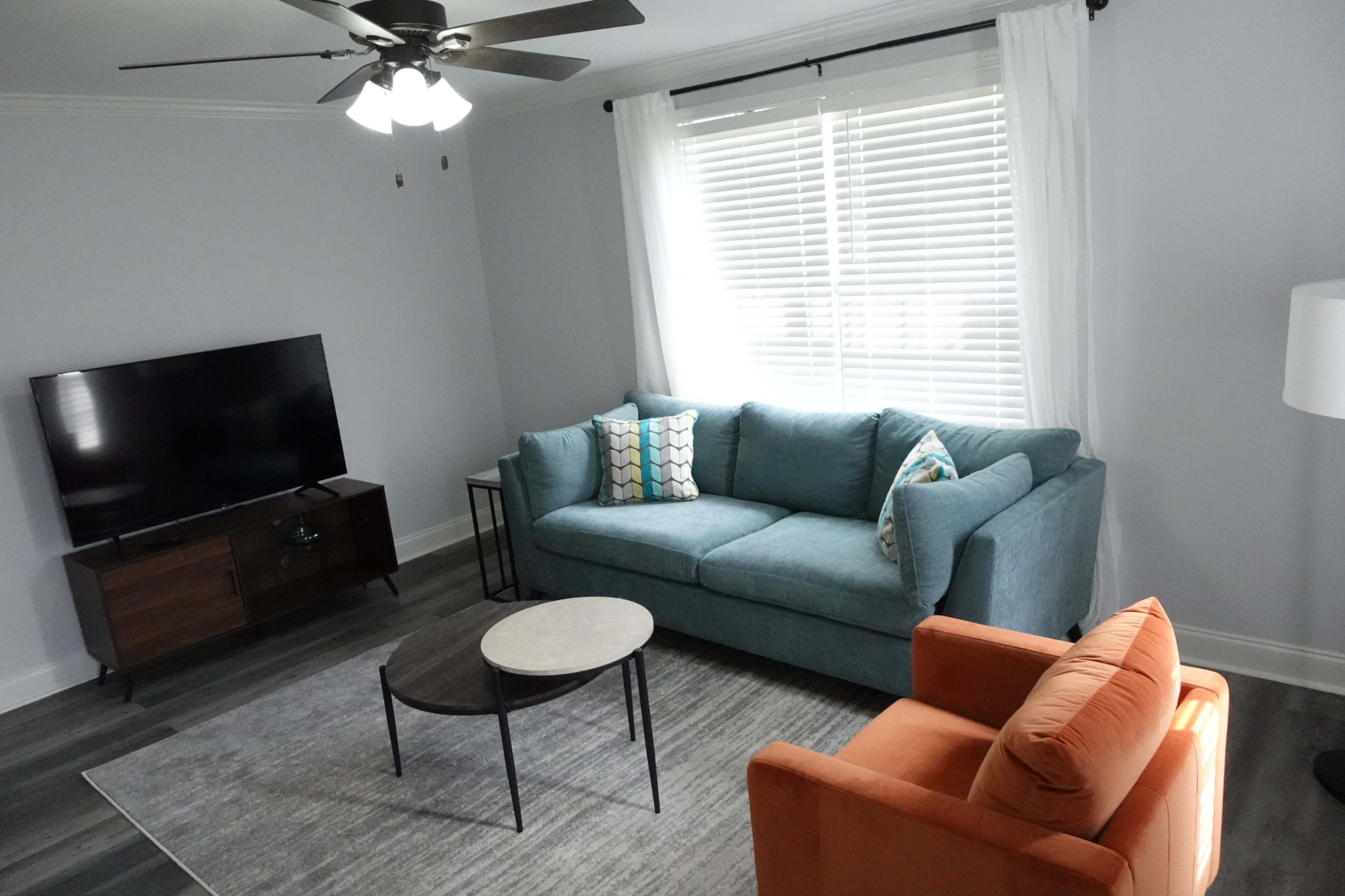 A living room with a teal sofa, an orange armchair, a round coffee table, and a television on a stand against the wall.