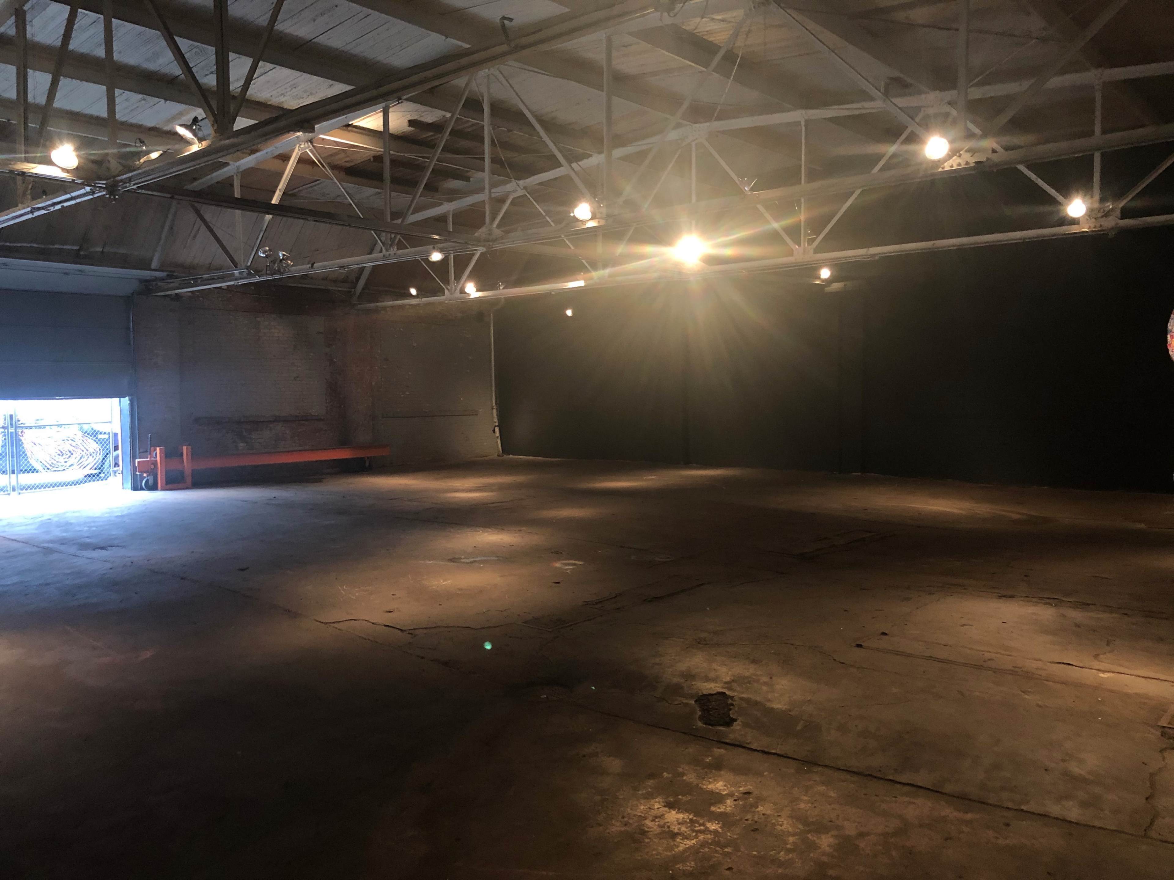 An empty industrial space with a concrete floor, exposed beams, and lighting fixtures, featuring a doorway that leads to an exterior area.