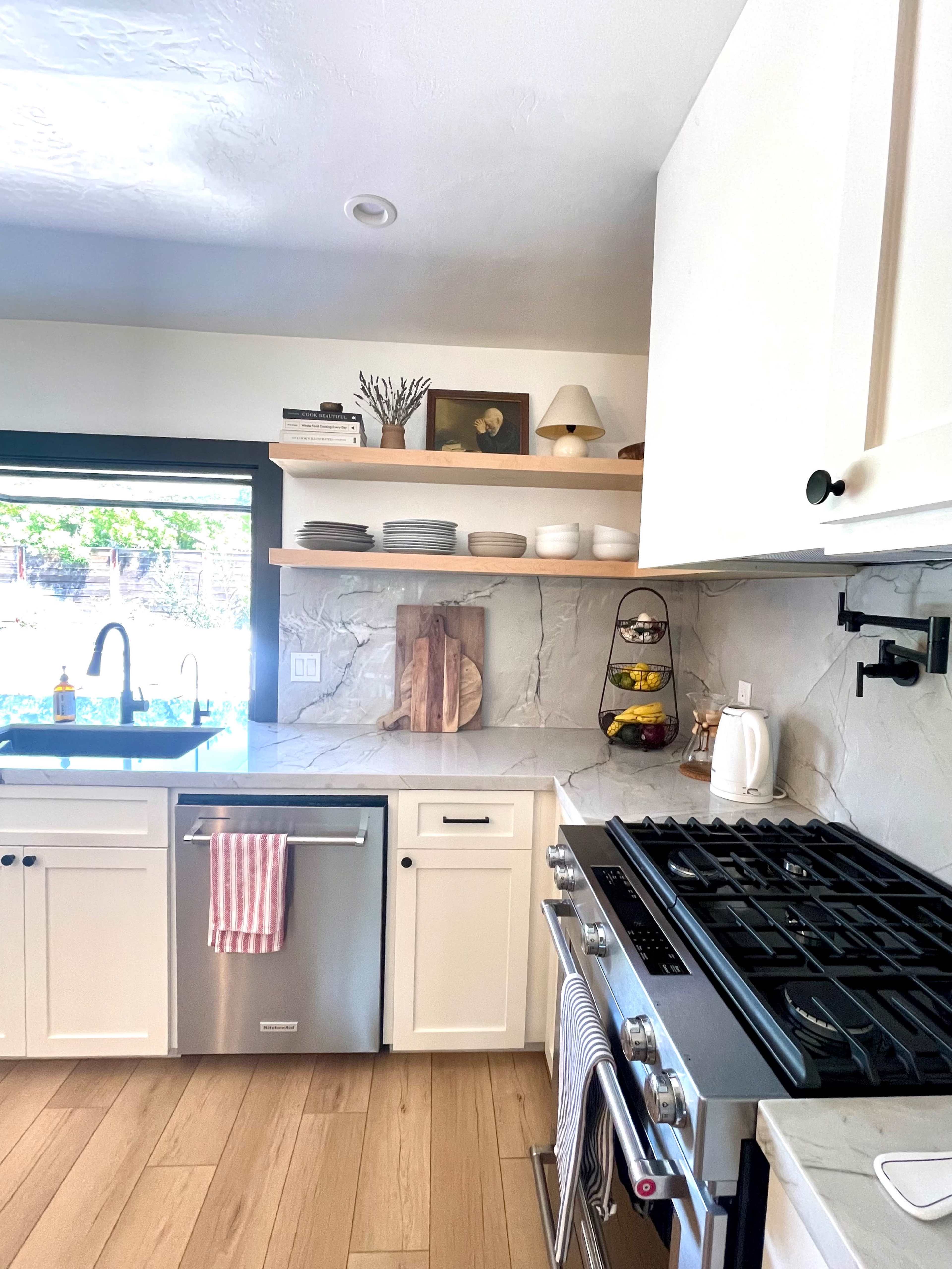 A modern kitchen with white cabinetry, a stainless steel dishwasher, and a gas stove, featuring open shelving above a counter with decorative items.