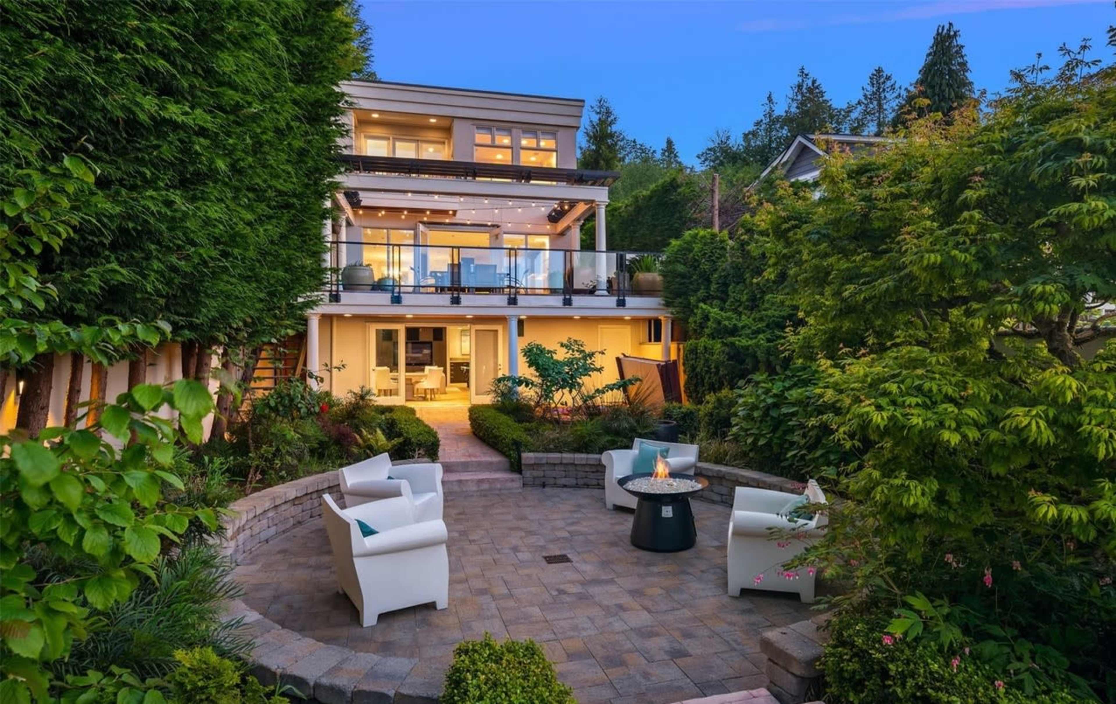 A modern two-story house surrounded by lush greenery and a circular patio area with white chairs and a fire pit.