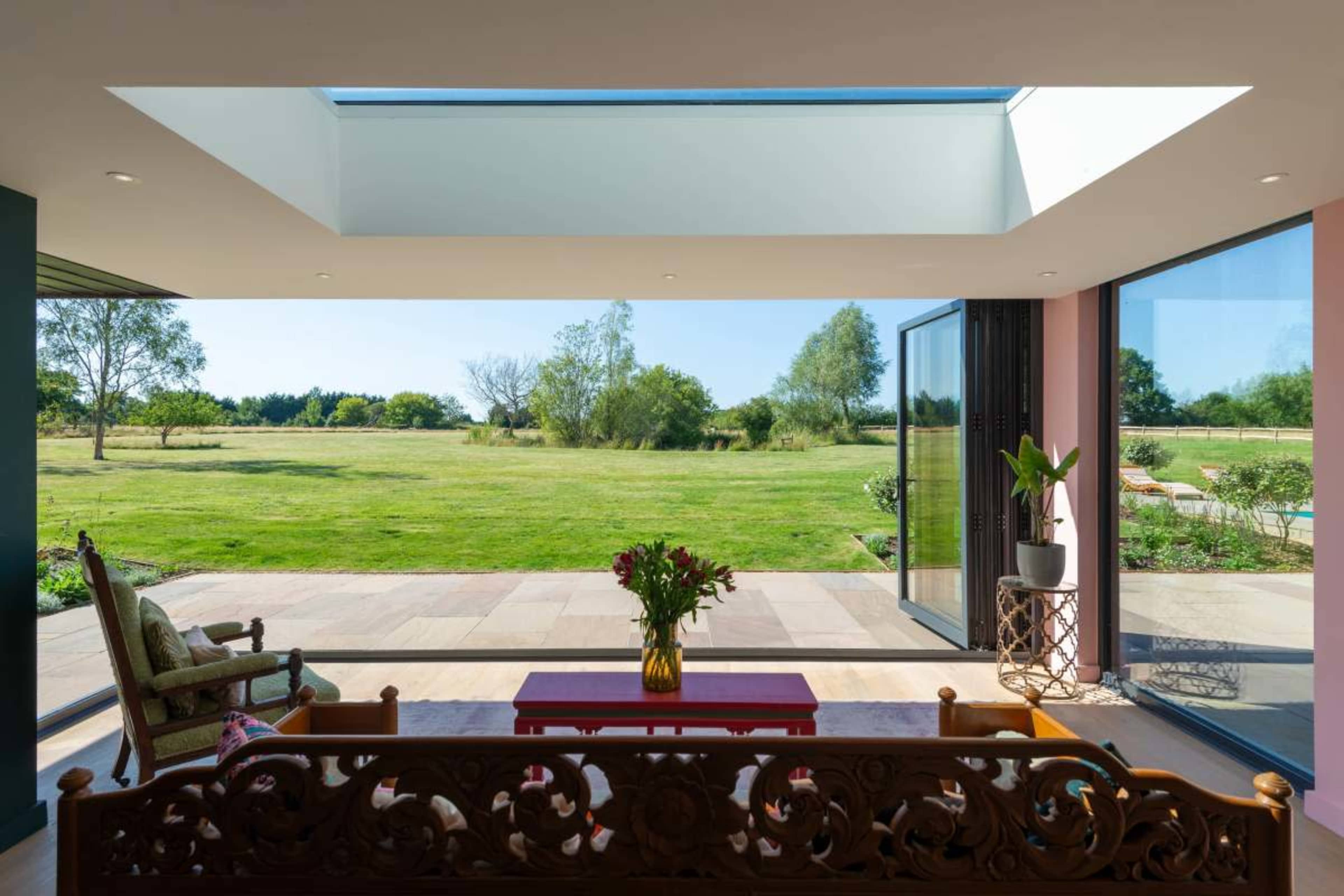 A spacious living area features large glass doors that open to a green landscape, with a wooden bench and a vase of flowers in the foreground.