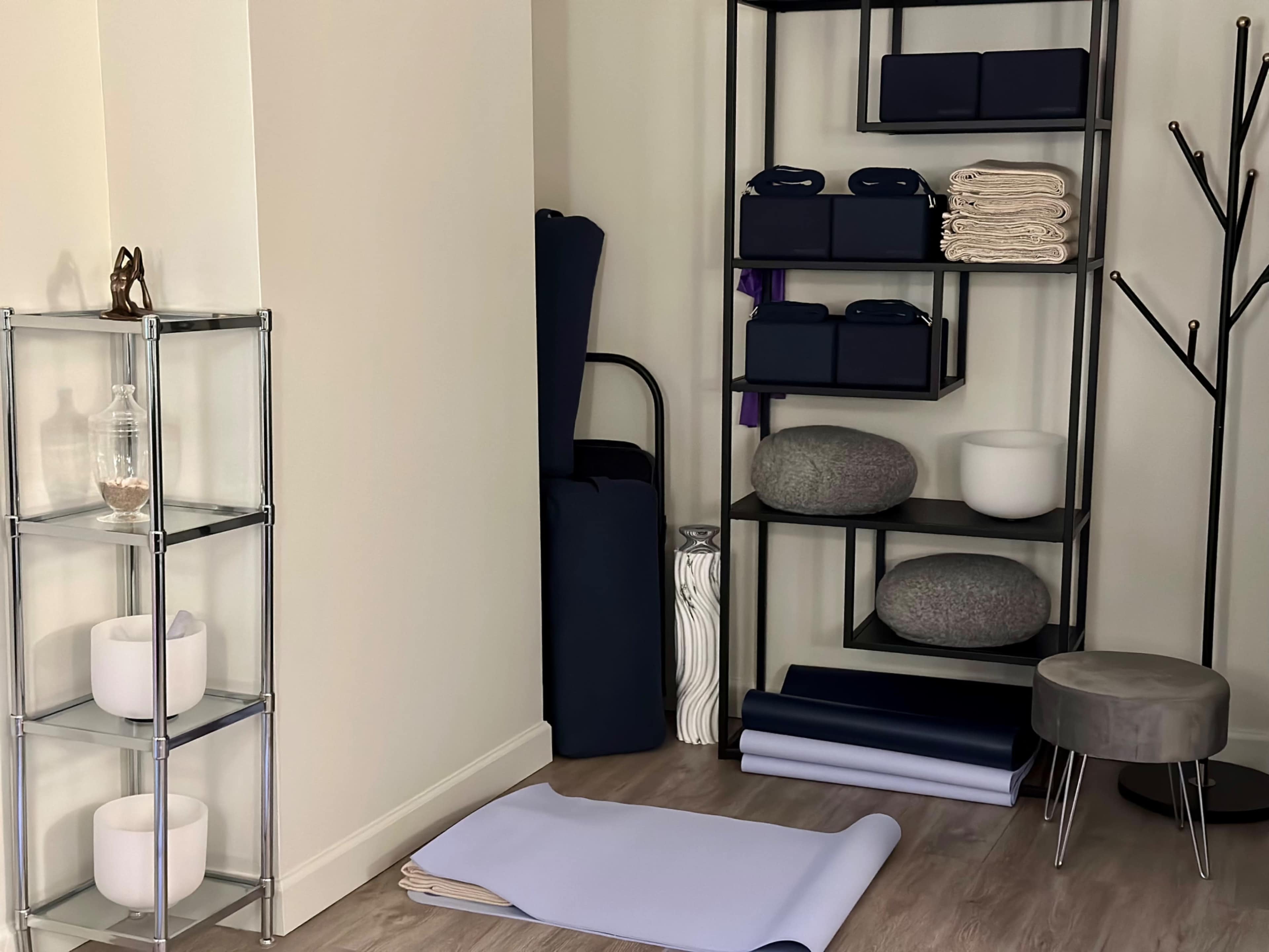 The image shows a serene yoga space with a neatly arranged shelving unit containing towels and mats, alongside a glass display unit holding decorative items.