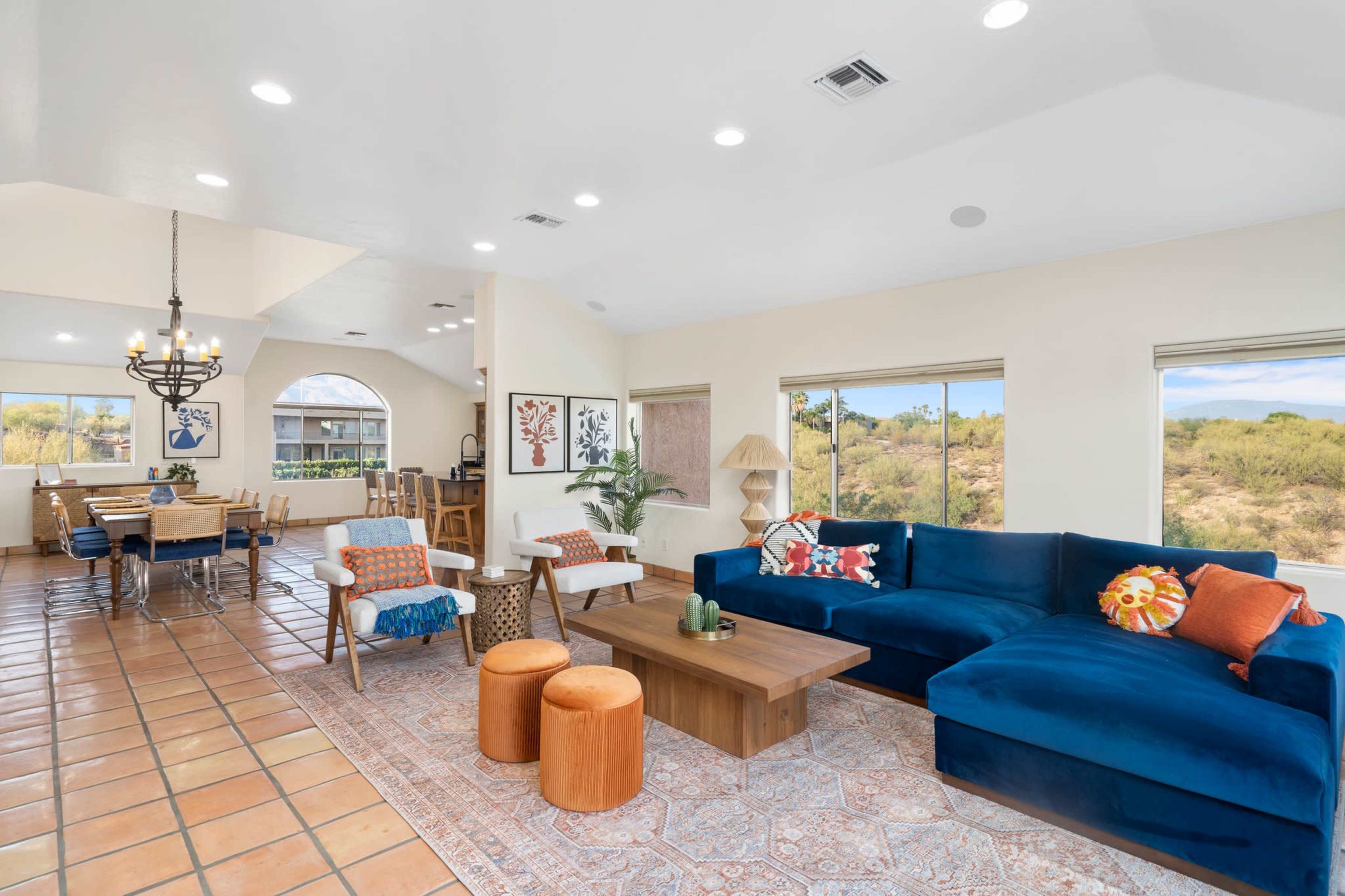 A bright, open living area features a blue sectional couch, wooden coffee table, and dining space that leads to large windows showcasing a desert landscape.