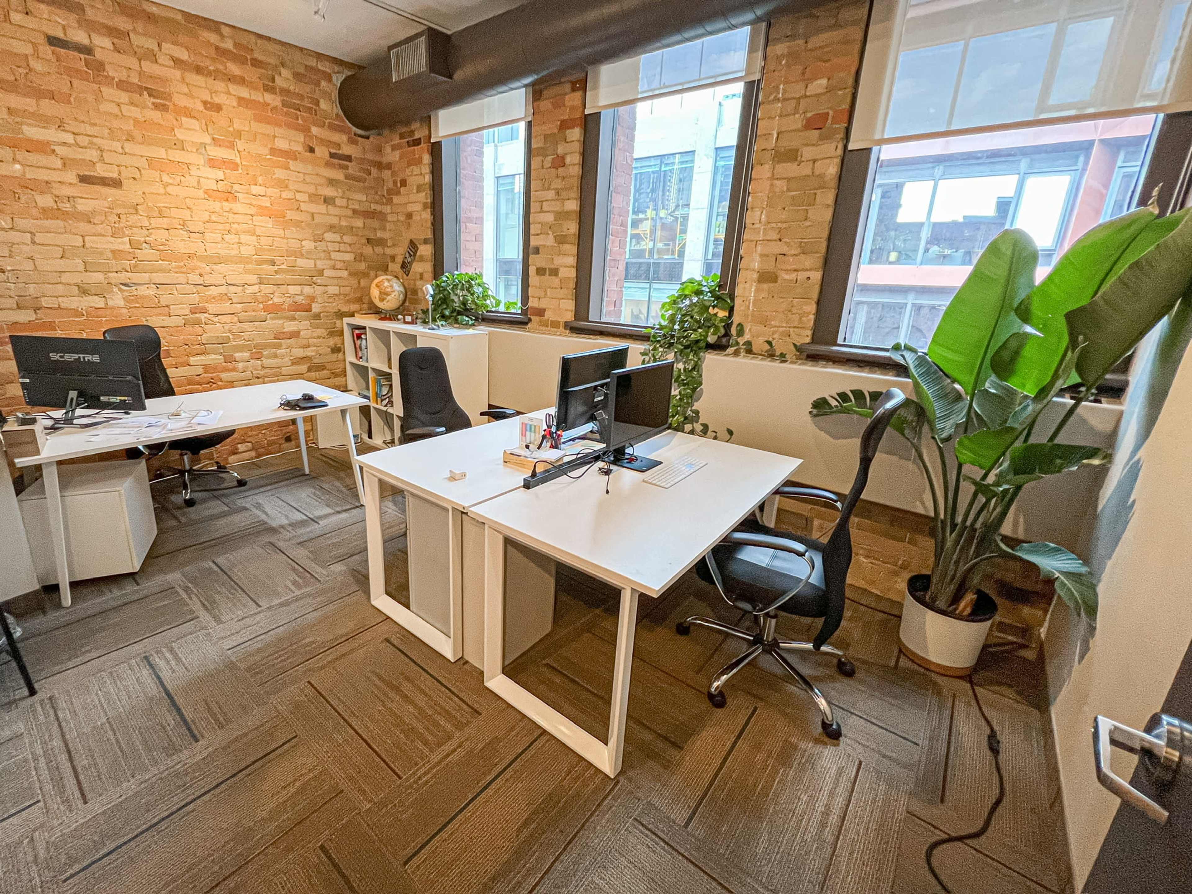 A modern office space with two desks, plants, large windows, and exposed brick walls.