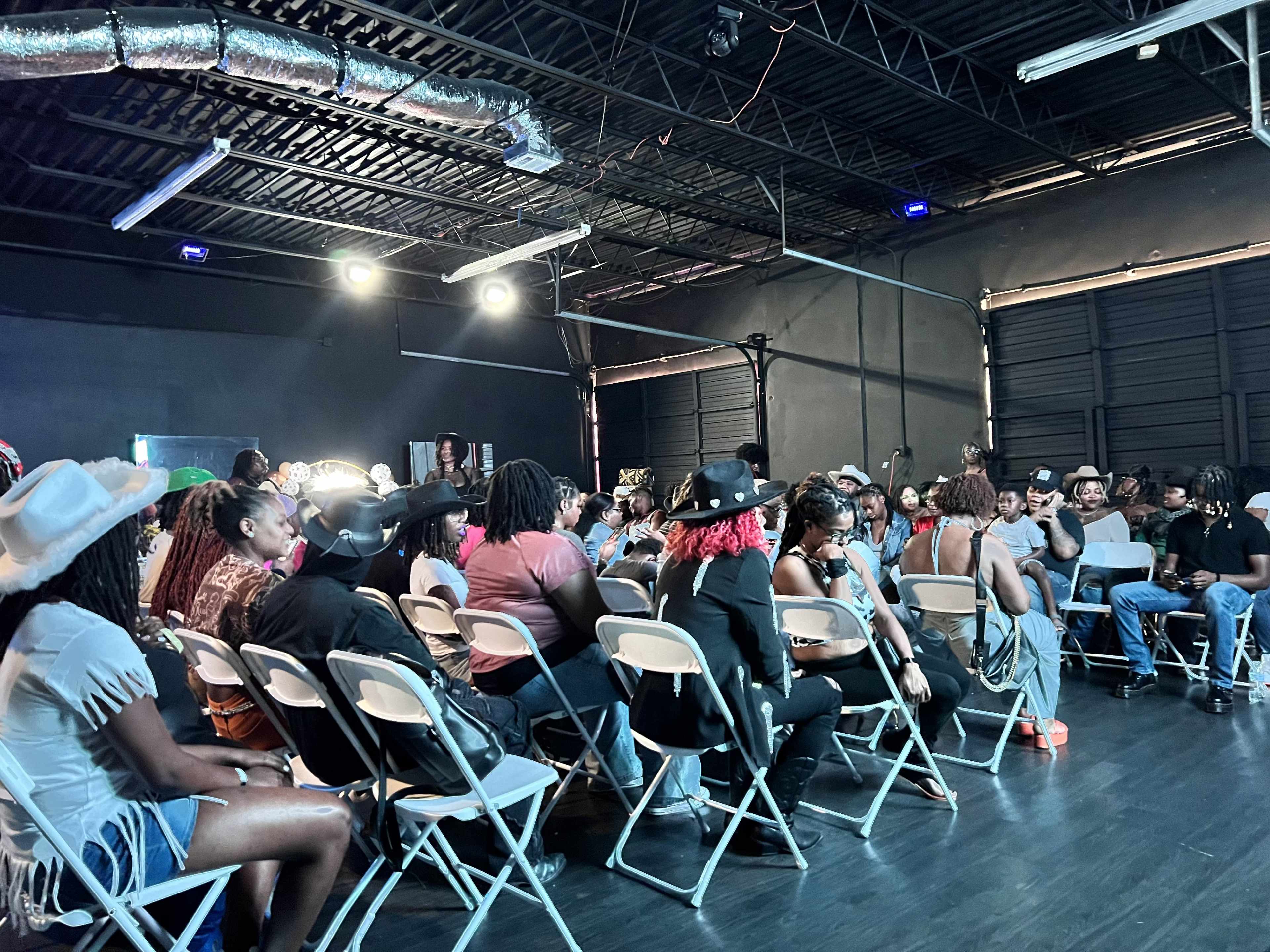 A diverse audience sits in folding chairs, facing a performance stage in an industrial-style venue.