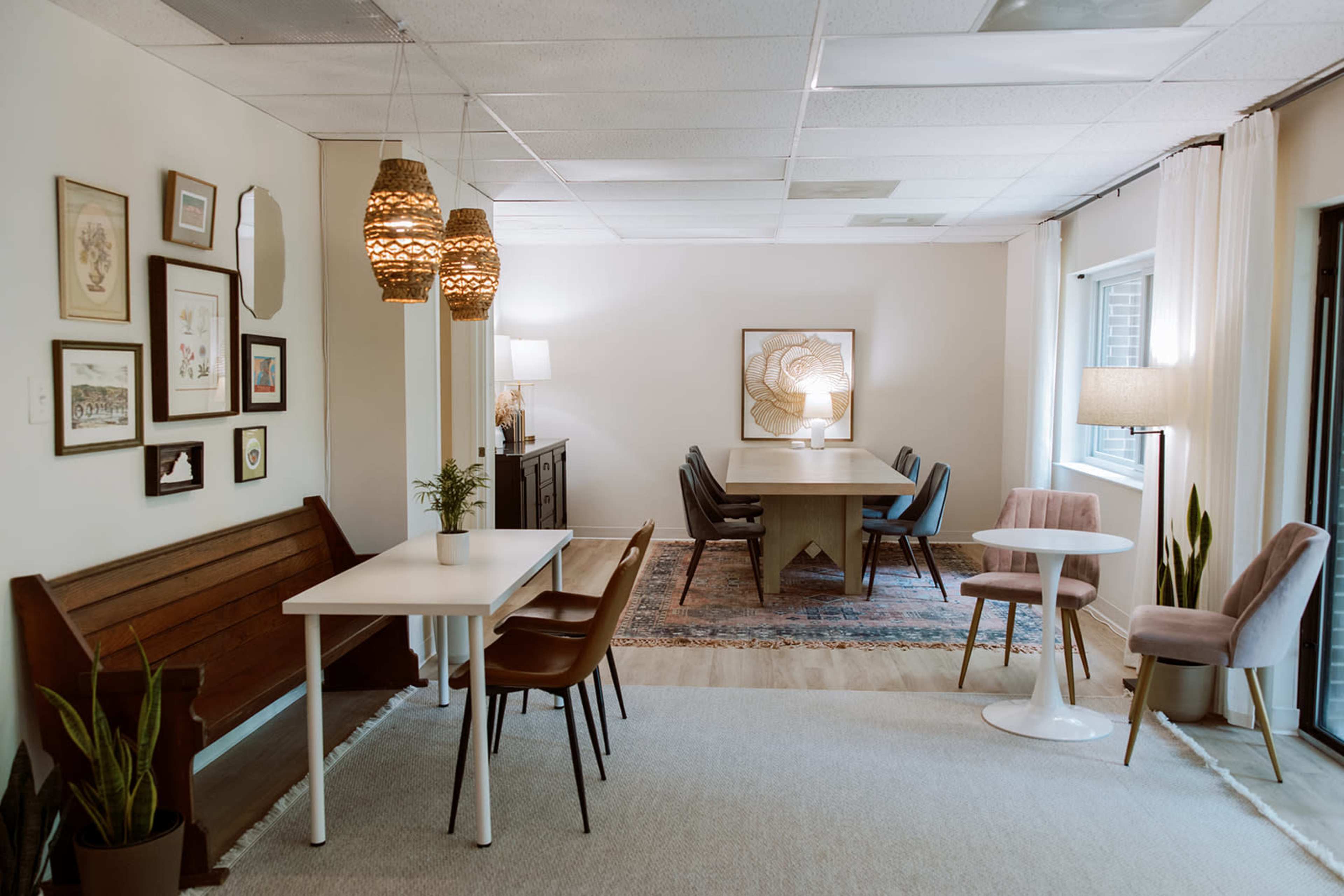 The image shows a modern interior space featuring a combination of seating areas, including a dining table with chairs, a small side table, and a decorative wall with artwork and plants.