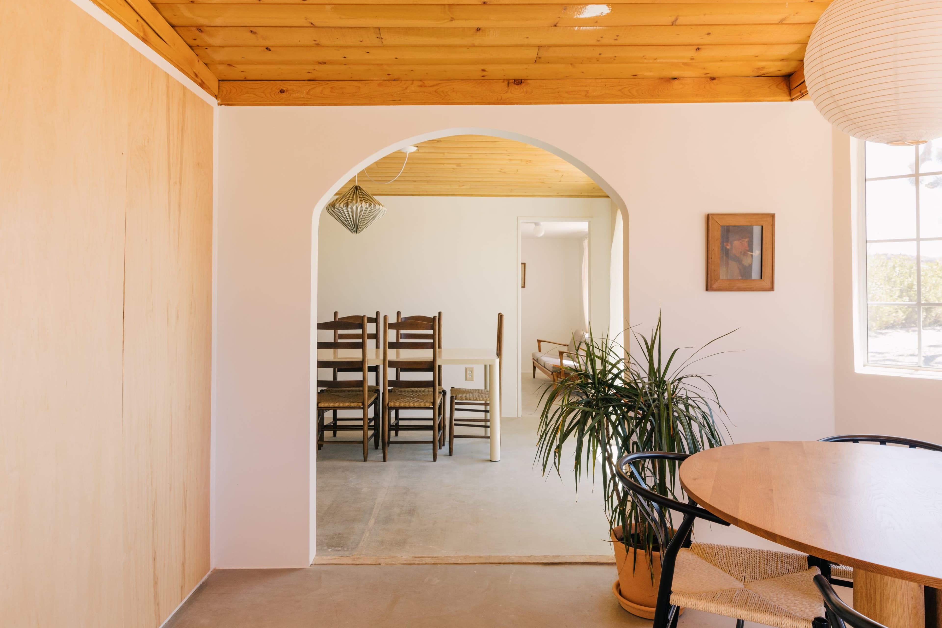 The image shows a room with a wooden ceiling, an arched doorway leading to a space with stacked chairs, and a potted plant next to a round table.