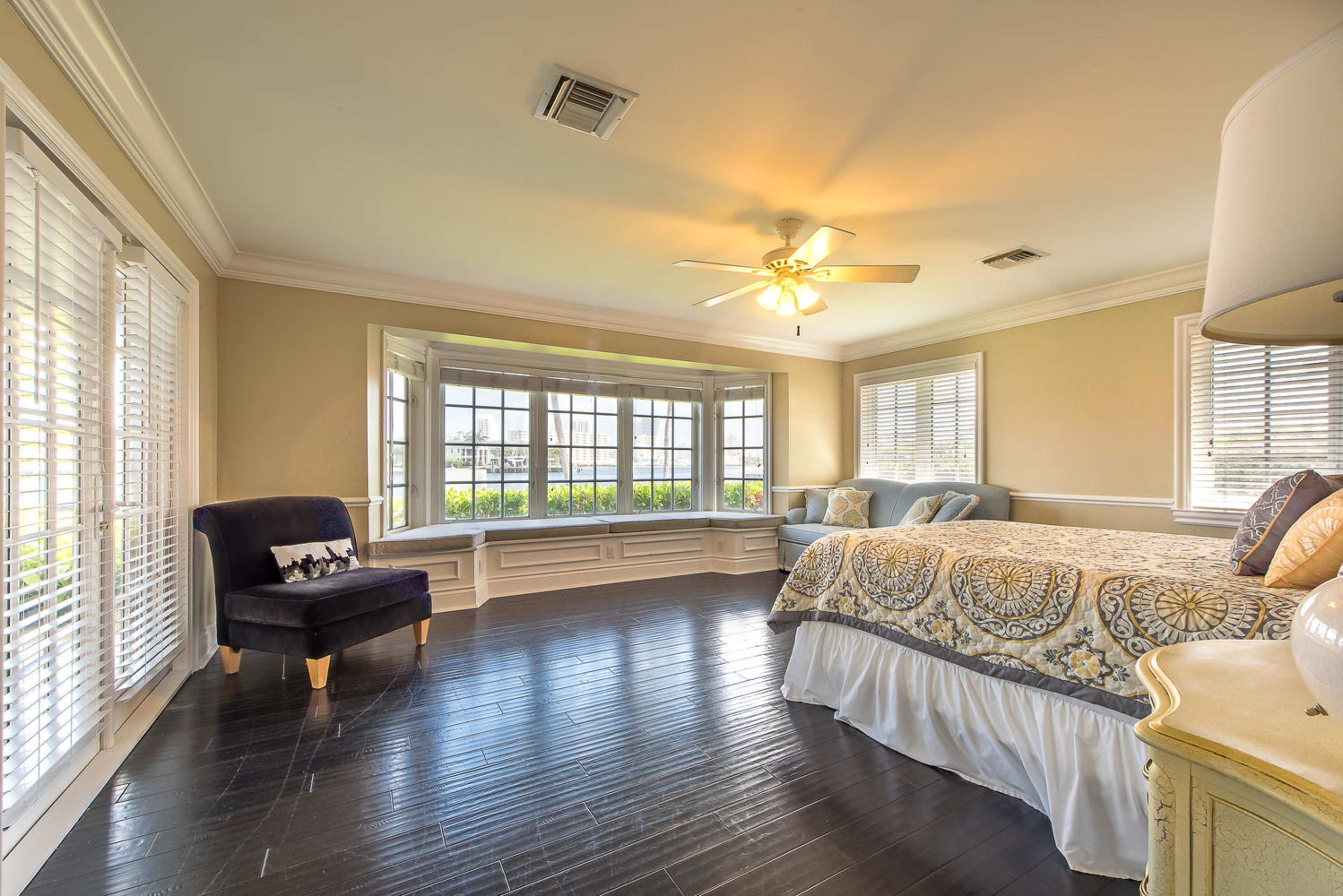 A spacious bedroom features a large window seat with multiple windows, hardwood flooring, and a bed with decorative bedding.