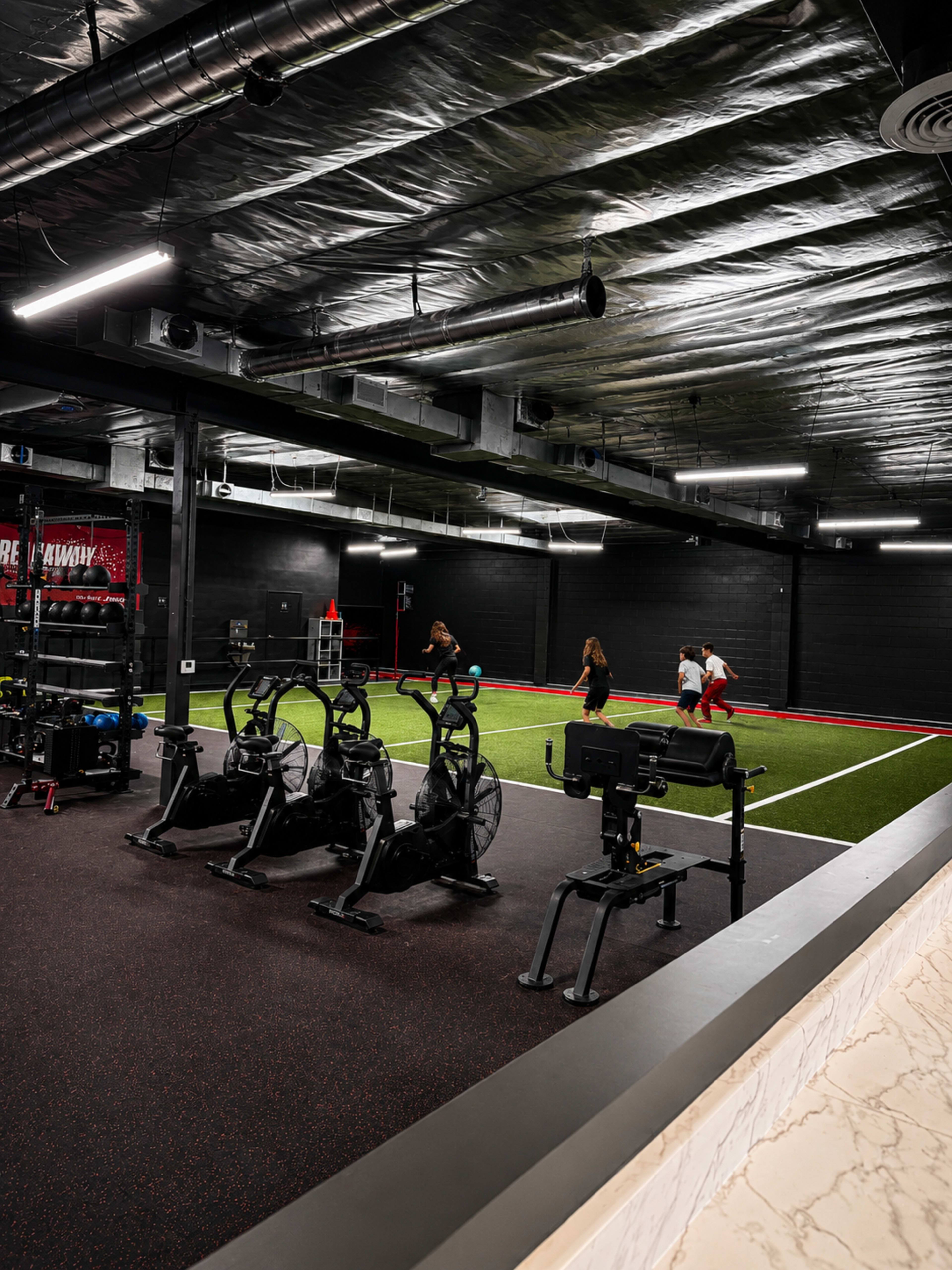 The image shows a modern indoor gym space with exercise equipment, including stationary bikes and rowing machines, and individuals engaging in workouts on a turf area.