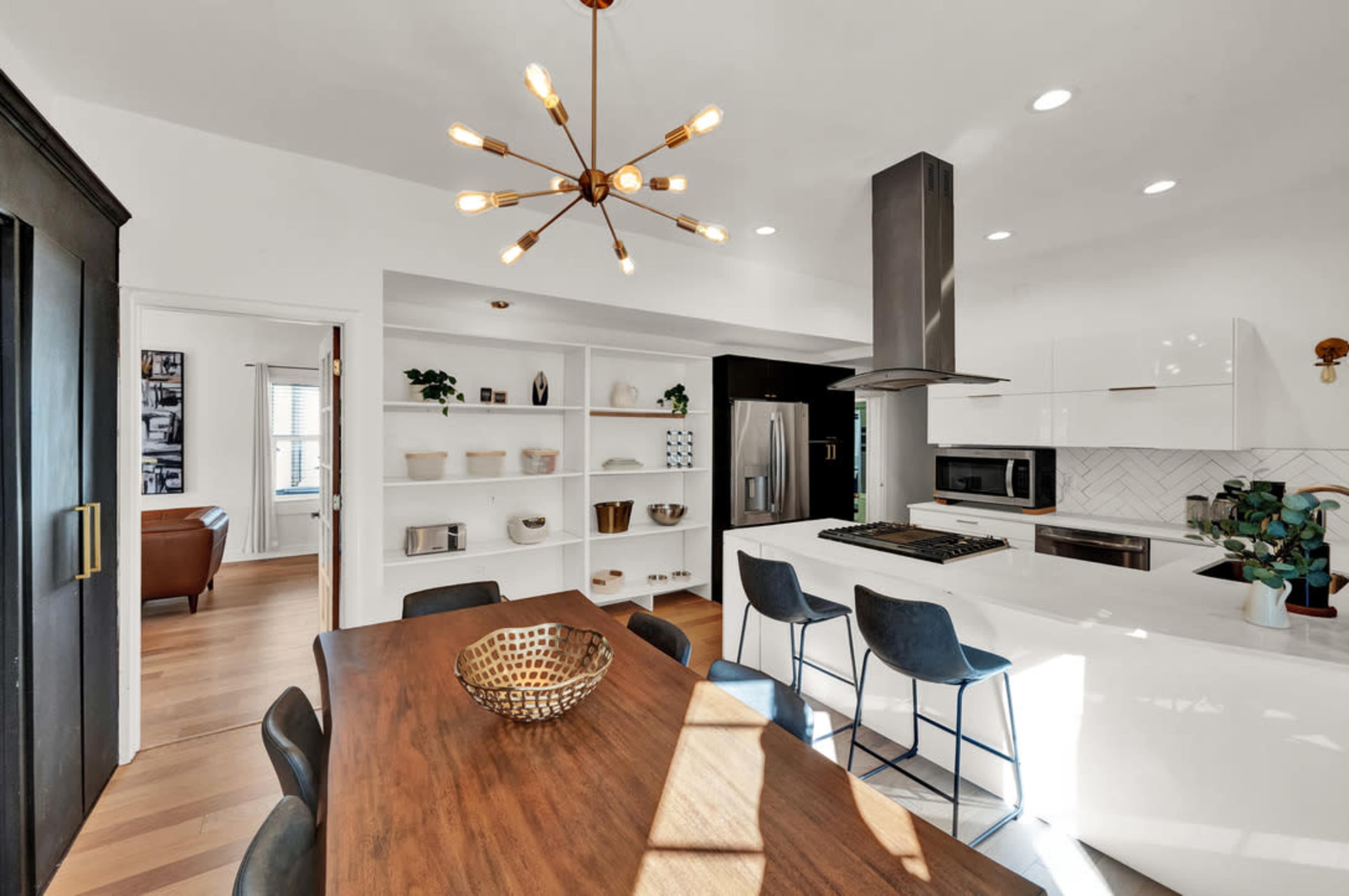 A modern kitchen with white cabinetry, a stainless steel hood, and a dining table set with a decorative bowl, illuminated by pendant lights.