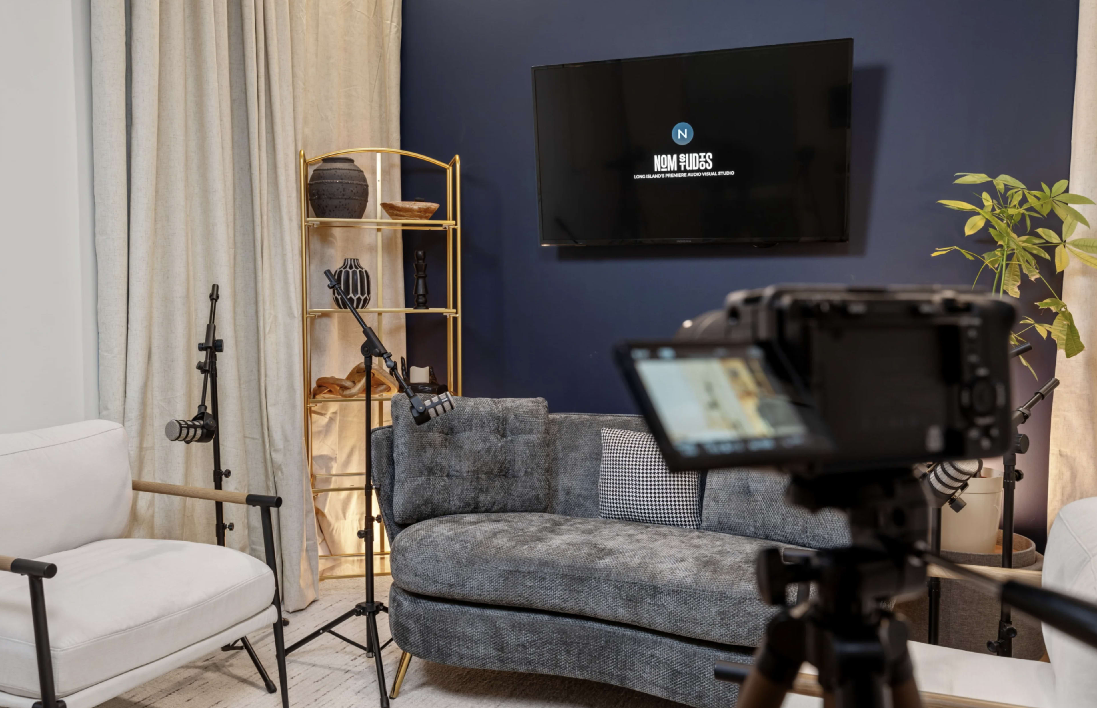 The image shows a modern living room setup with a gray couch, two light-colored chairs, and a television mounted on a dark blue wall, along with microphones and a plant in the background.