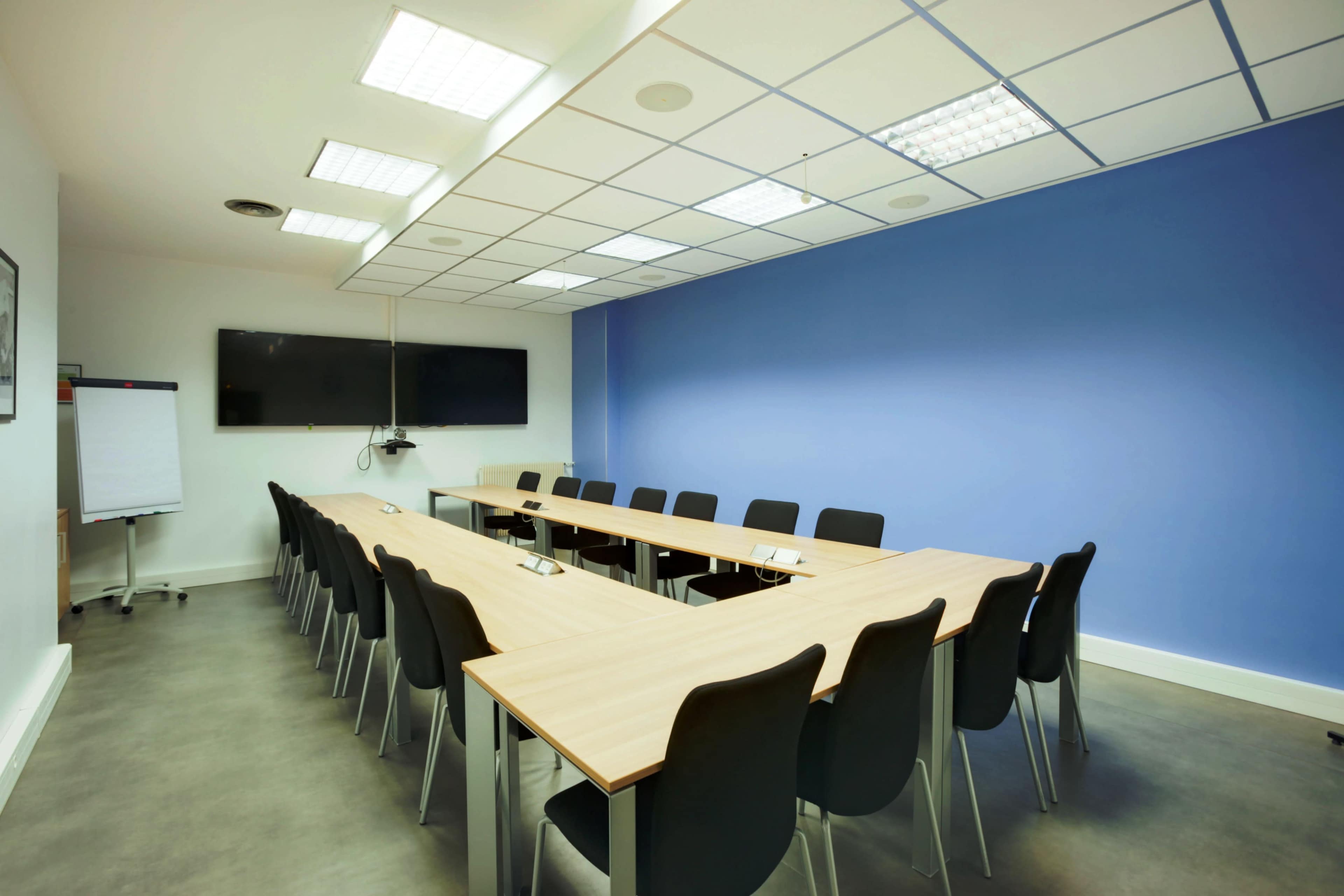 A conference room features a large rectangular table surrounded by black chairs, with a wall displaying a blue color and a television mounted above it.