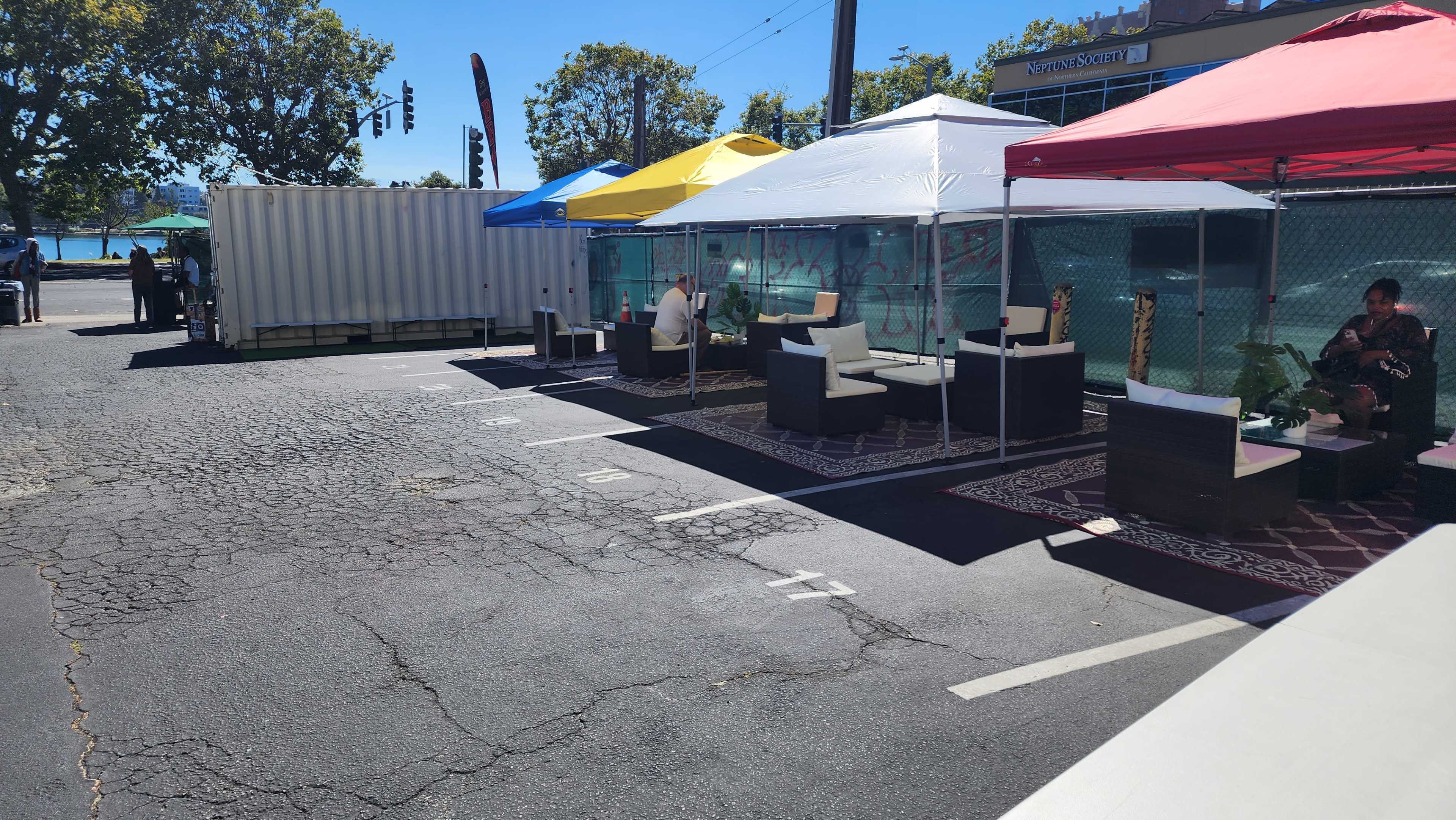 A row of tents with colorful canopies is set up in a parking lot, featuring seating areas and a shipping container in the background.