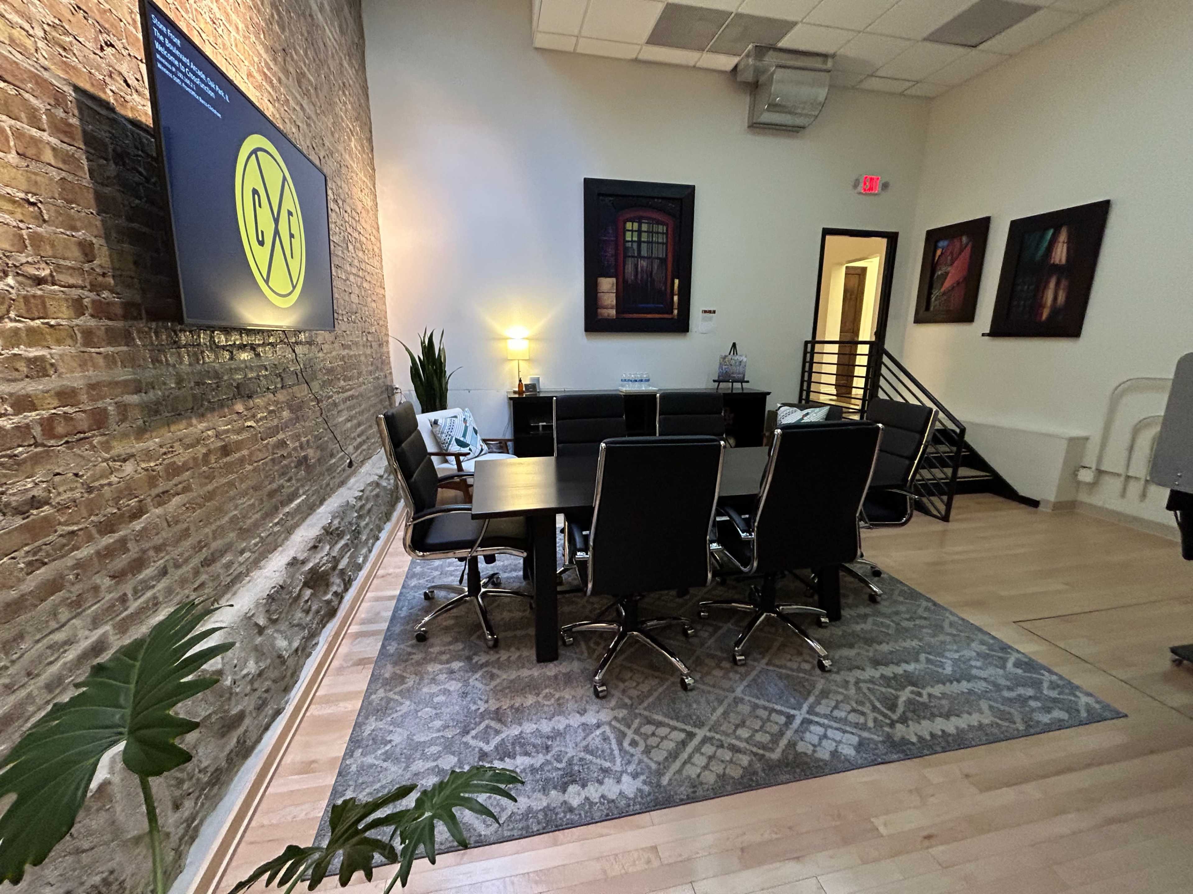 A modern conference room features a large table surrounded by black leather chairs, set against a brick wall with a television display and plants.