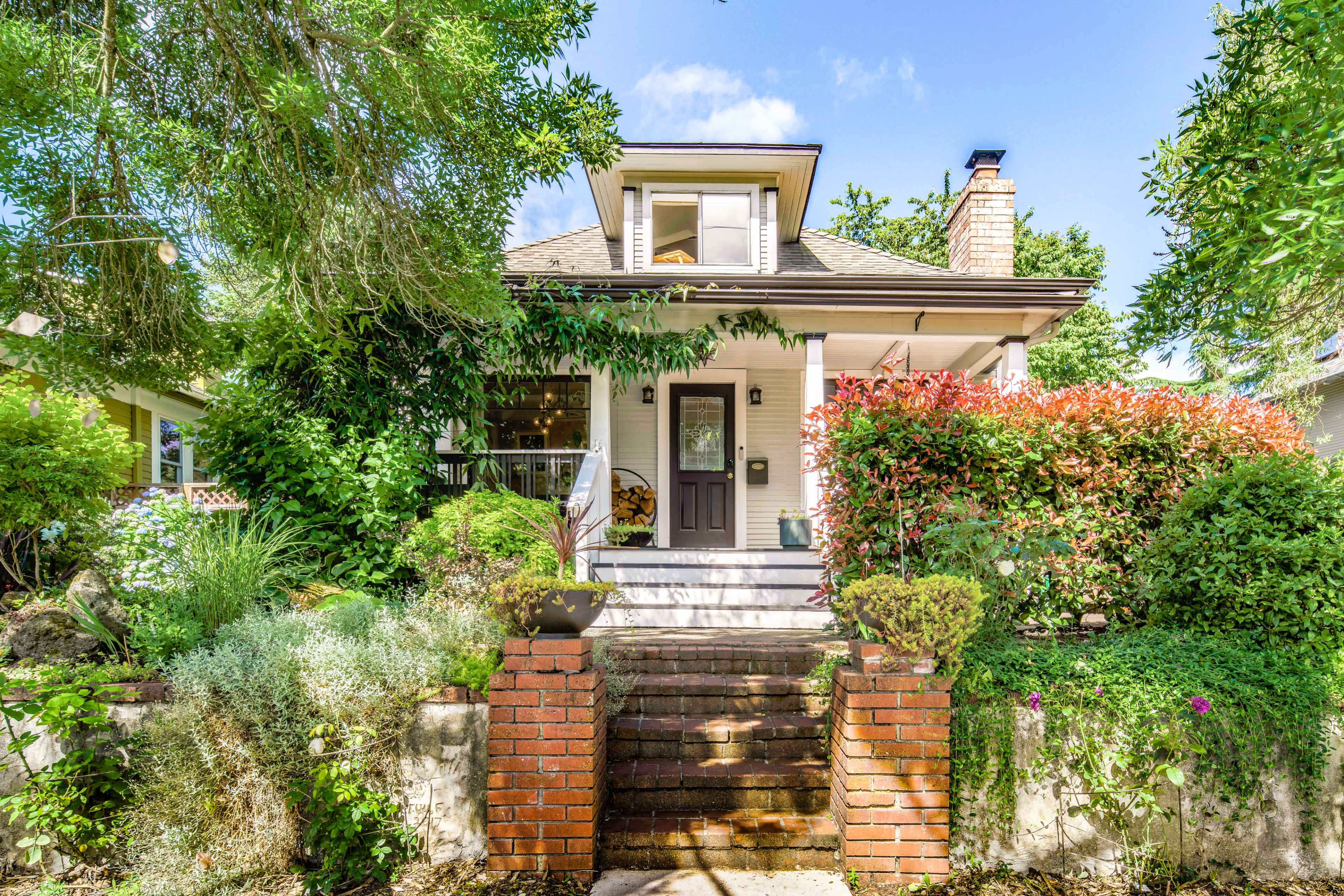 A two-story house with a sloped roof is surrounded by vibrant green and flowering plants, featuring a front staircase leading to the entrance.