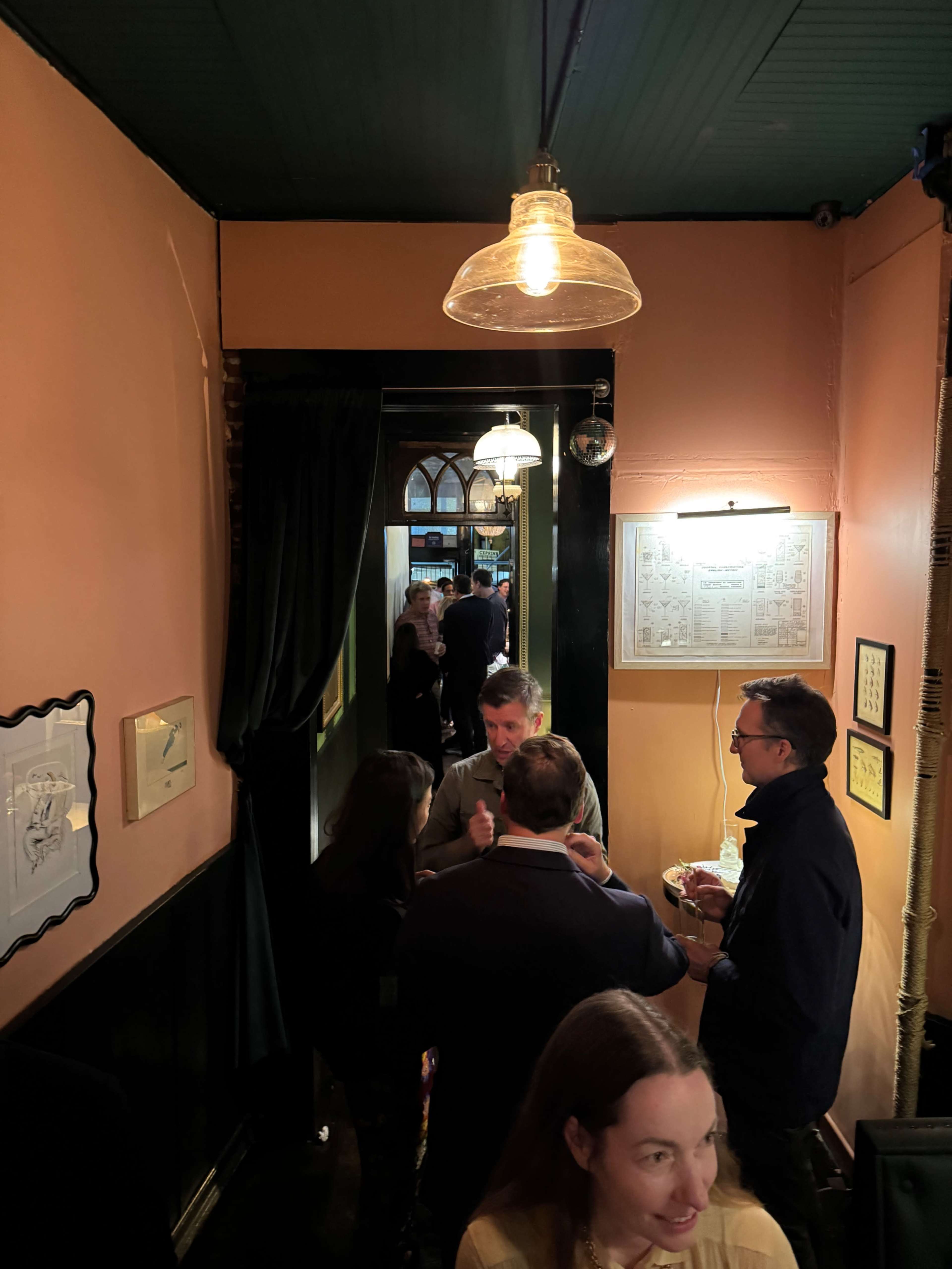 A narrow hallway in a bar features people socializing, with warm lighting and decorative elements on the walls.