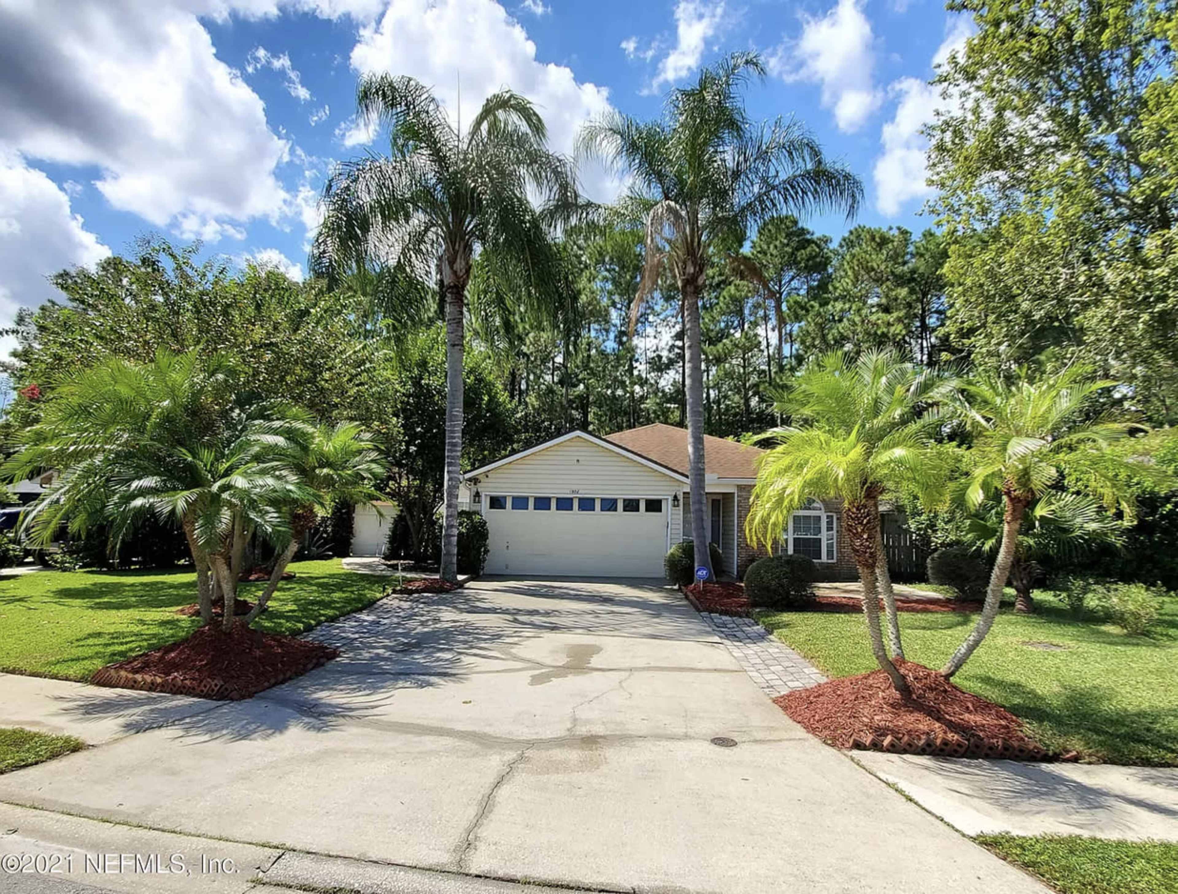 A single-story house with a driveway is surrounded by palm trees and landscaped greenery.