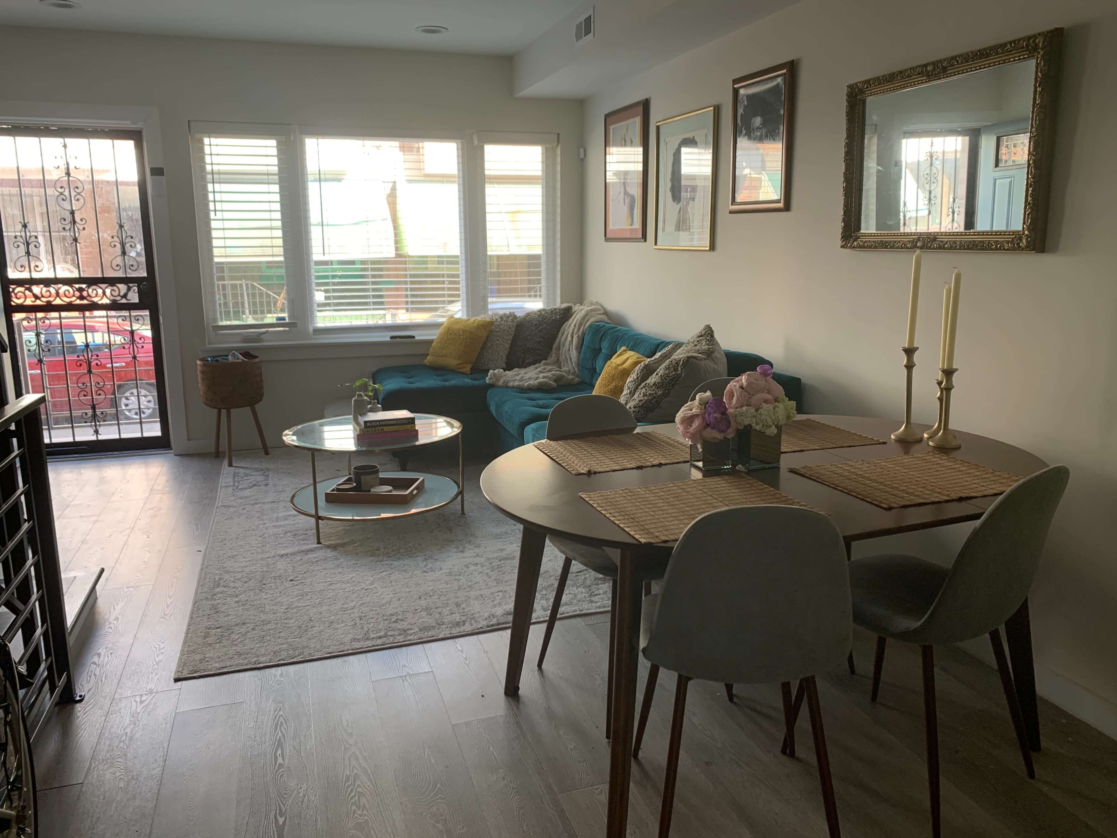 A cozy living area featuring a blue couch, a round coffee table, and a dining table set for two, with light streaming through window blinds.