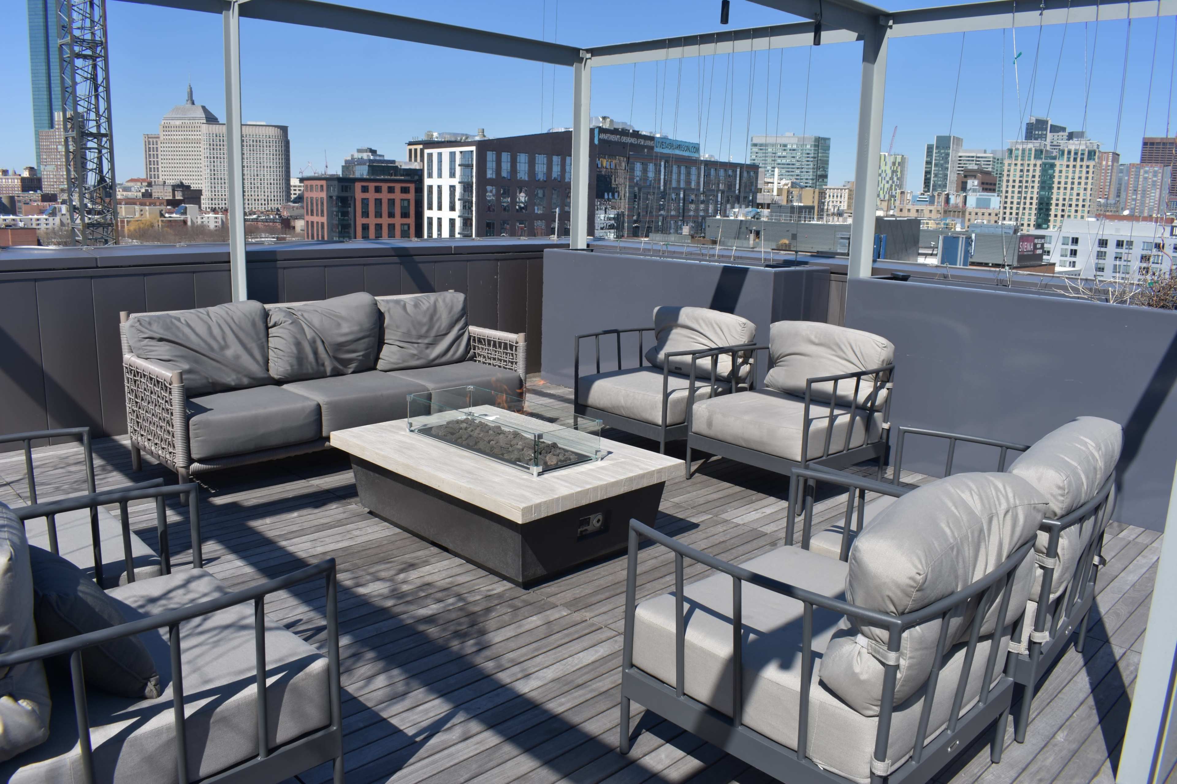 A rooftop lounge area with gray seating arrangements surrounding a central fire table, overlooking a city skyline.