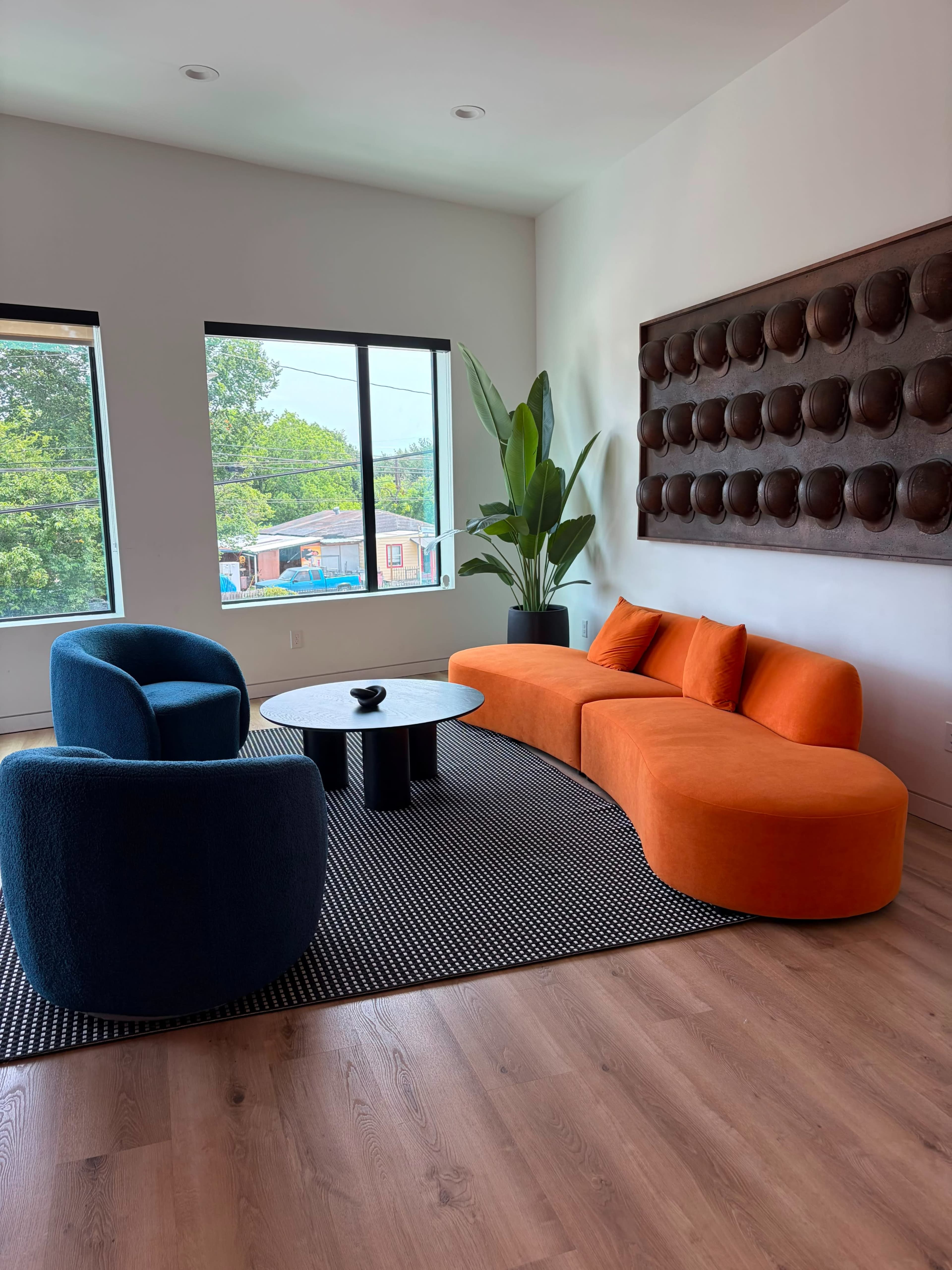 The room features a modern layout with an orange and blue seating arrangement, a round black table, and large windows overlooking greenery.