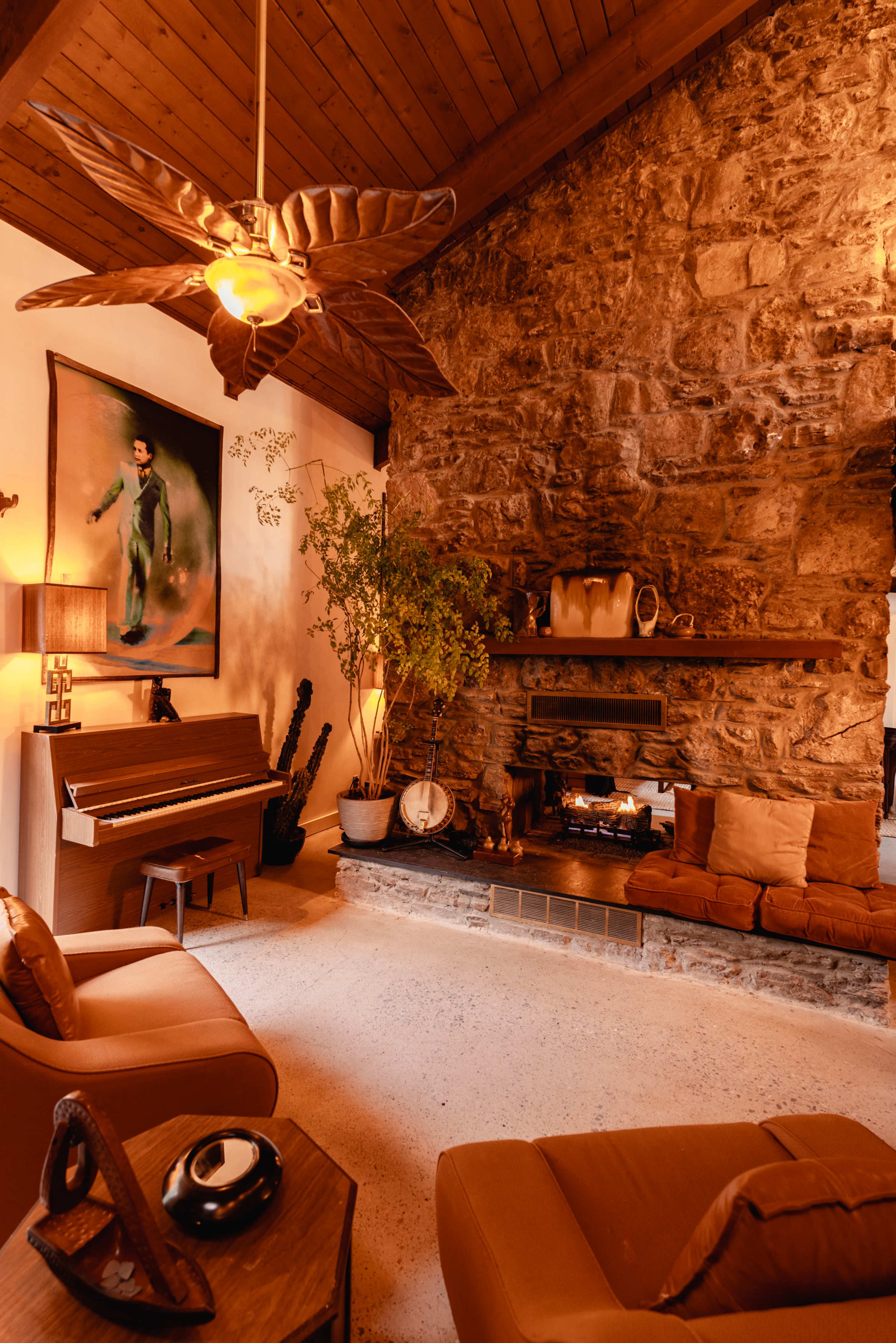 A cozy living room features a stone wall, a wooden ceiling, a piano, and a fireplace with a warm glow.