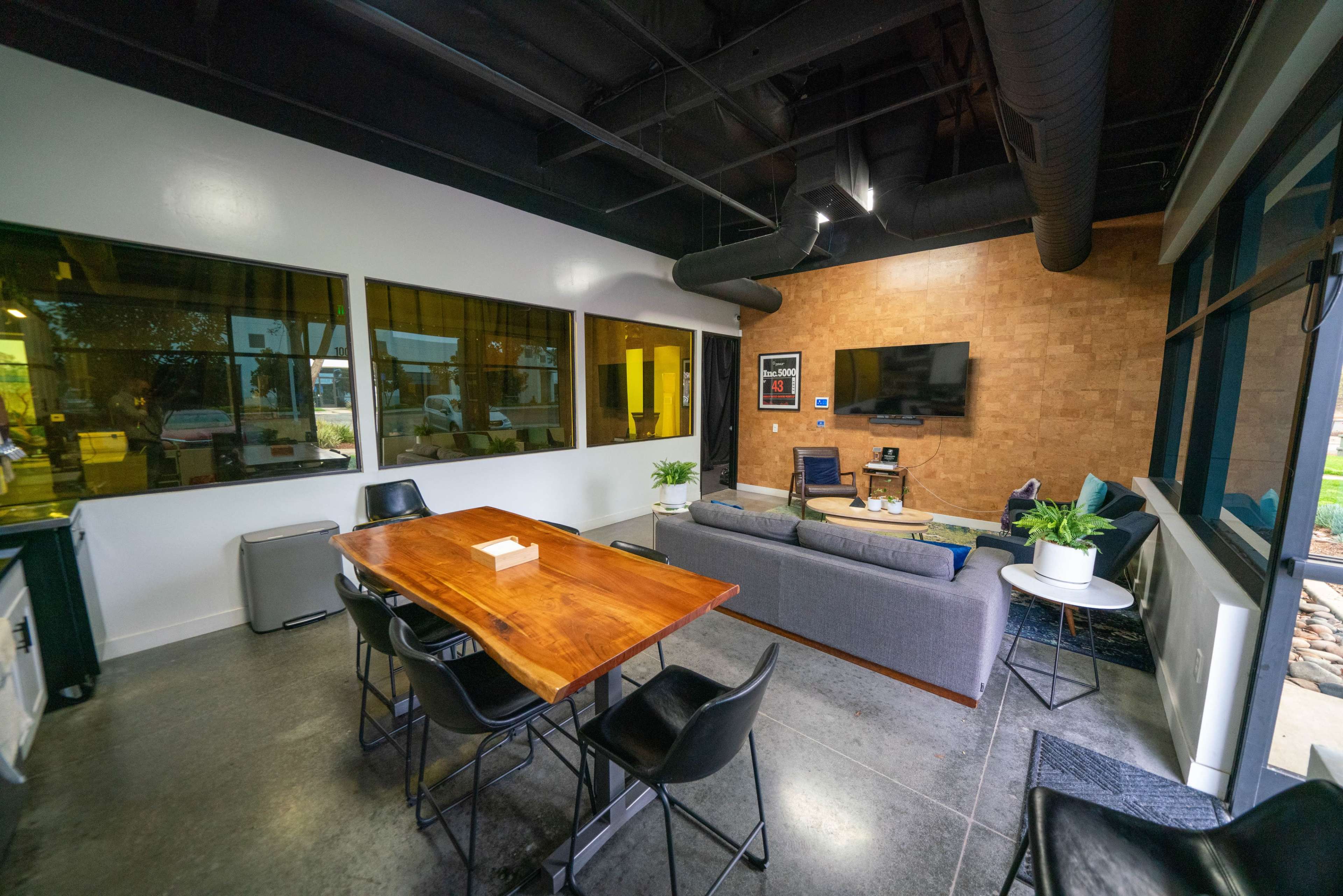 The image shows a modern interior space featuring a wooden dining table, a couch, a television mounted on the wall, and large windows that allow natural light to enter.