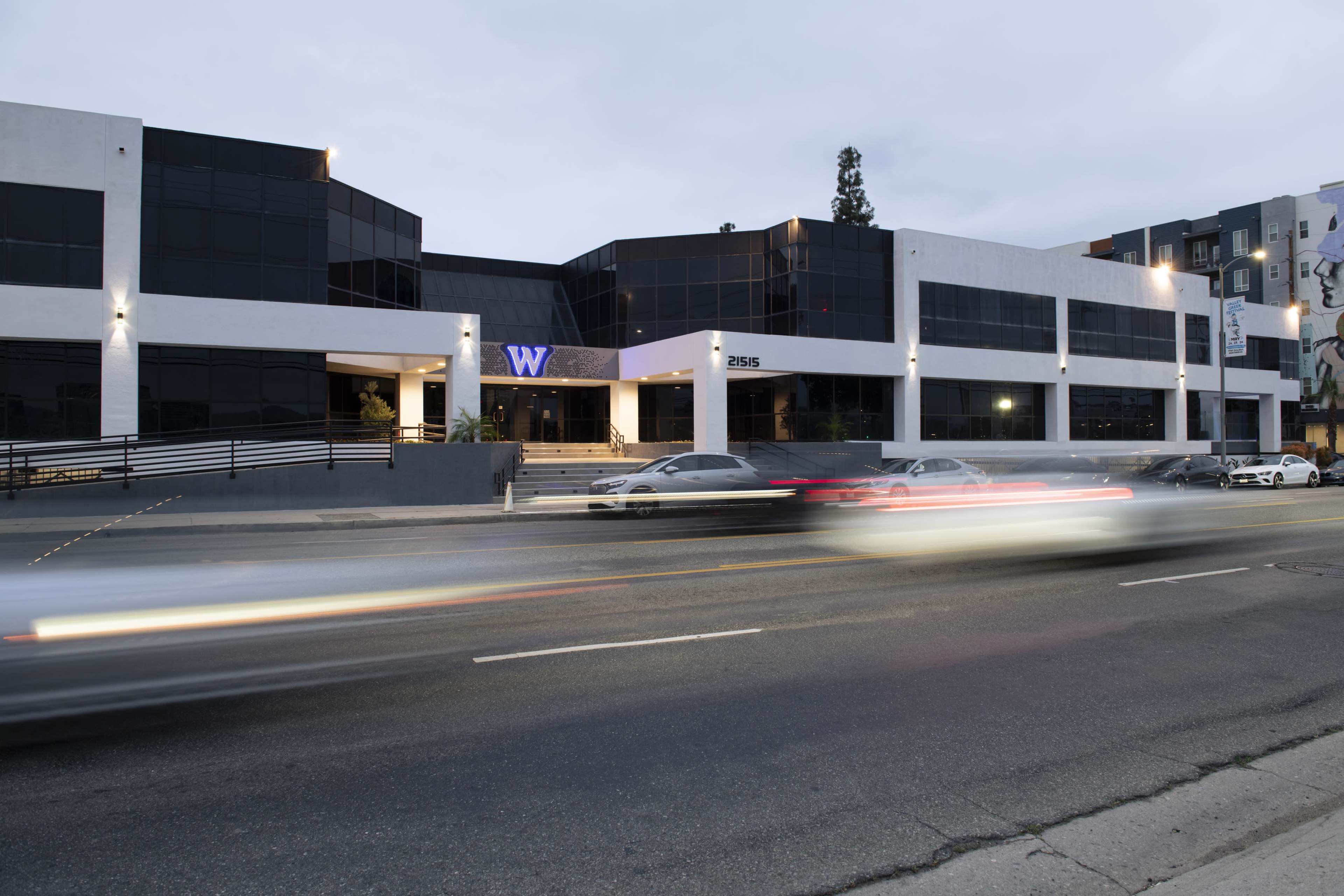 The image shows a modern, multi-story building with large glass windows, labeled with a "W," located along a bustling road, while cars pass by in motion.