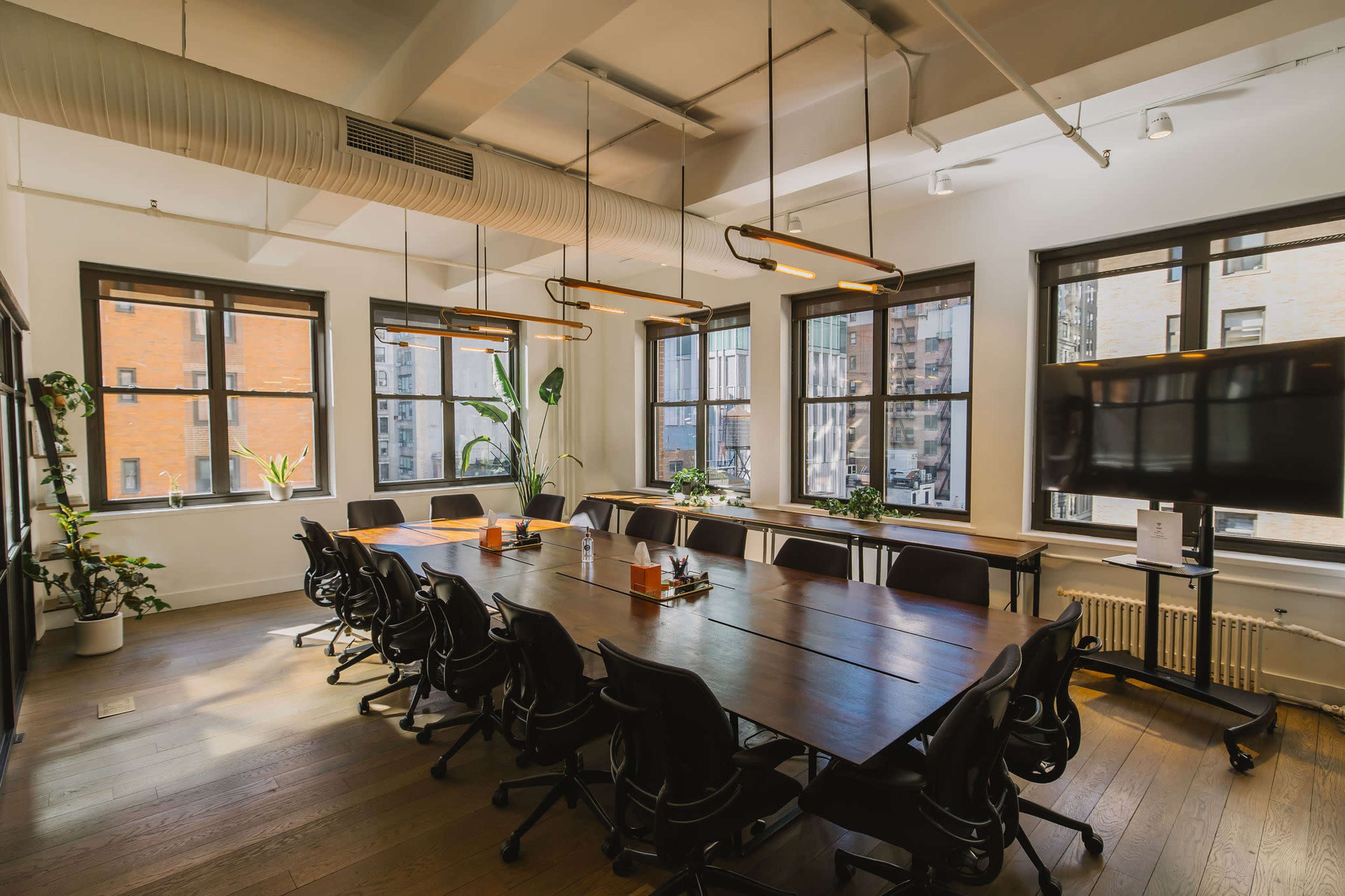 A modern conference room features a large wooden table surrounded by black office chairs, with multiple windows providing natural light and plants adding greenery.