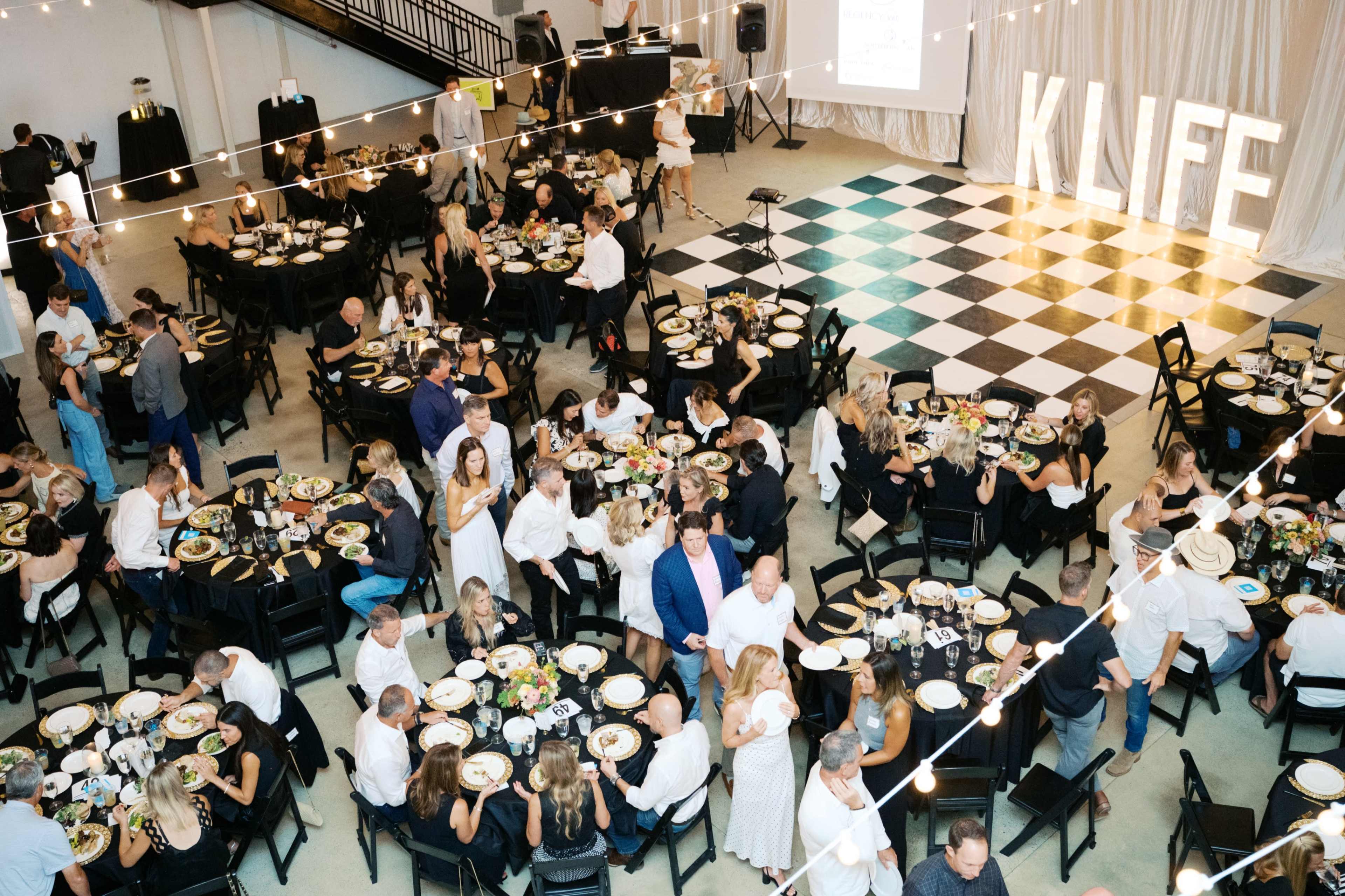 A large event space features several tables set for dinner, with guests seated and enjoying their meals under string lights, while a stage displays "KLIFE" in large letters.