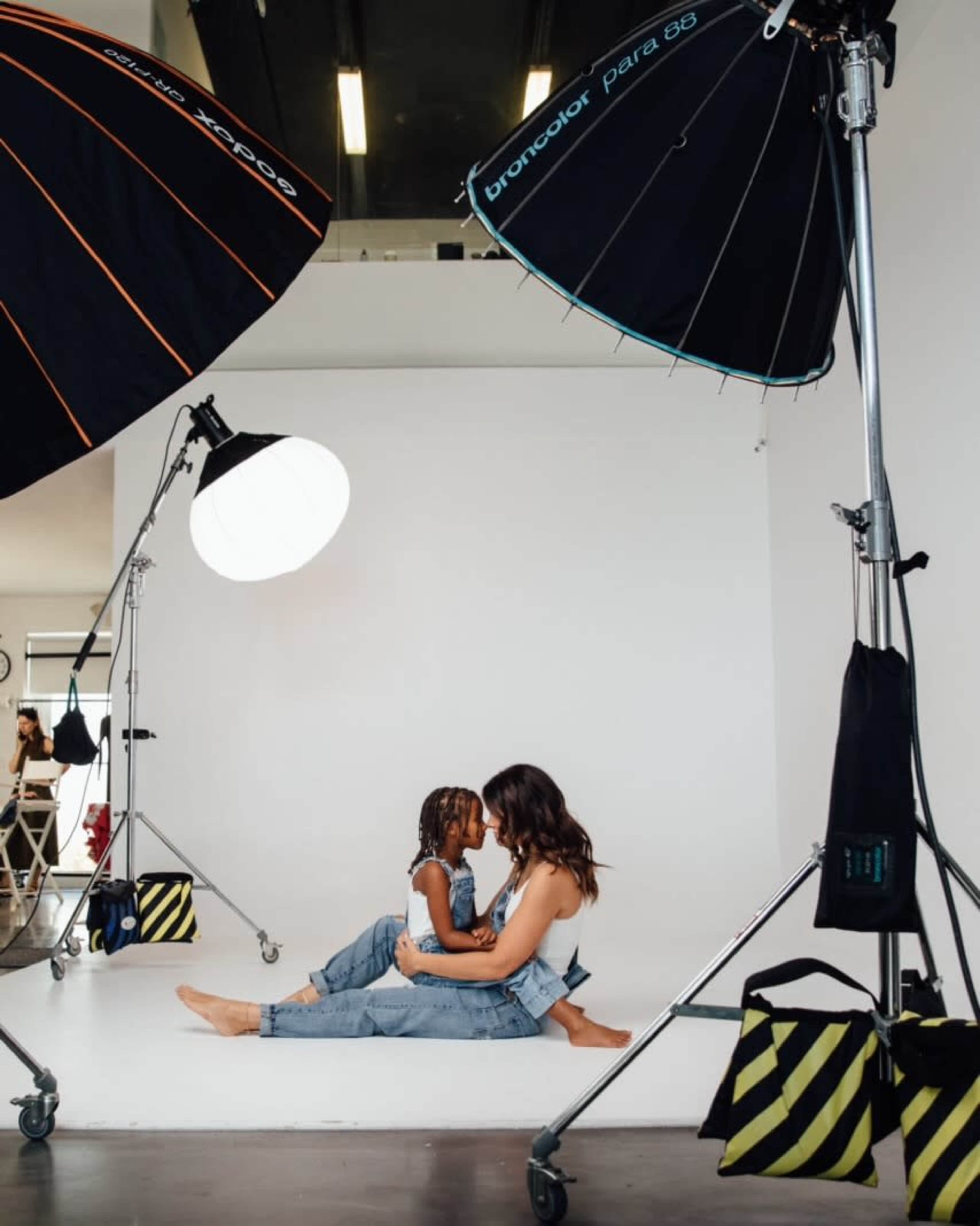 A woman and a child sit on the floor of a photography studio, surrounded by lighting equipment, as they face each other.