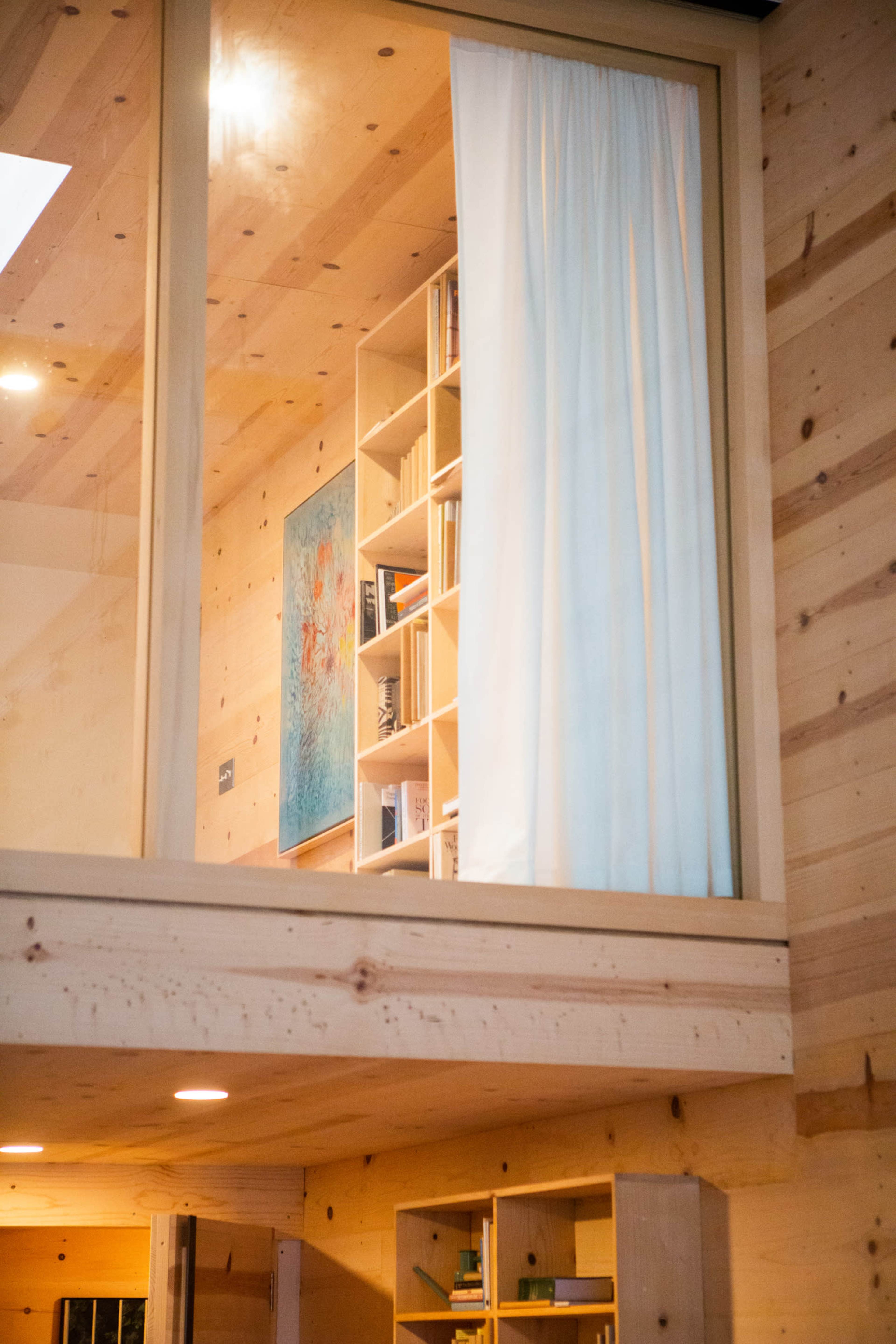 A wooden interior with a large window revealing a bookshelf and an art piece behind a curtain.
