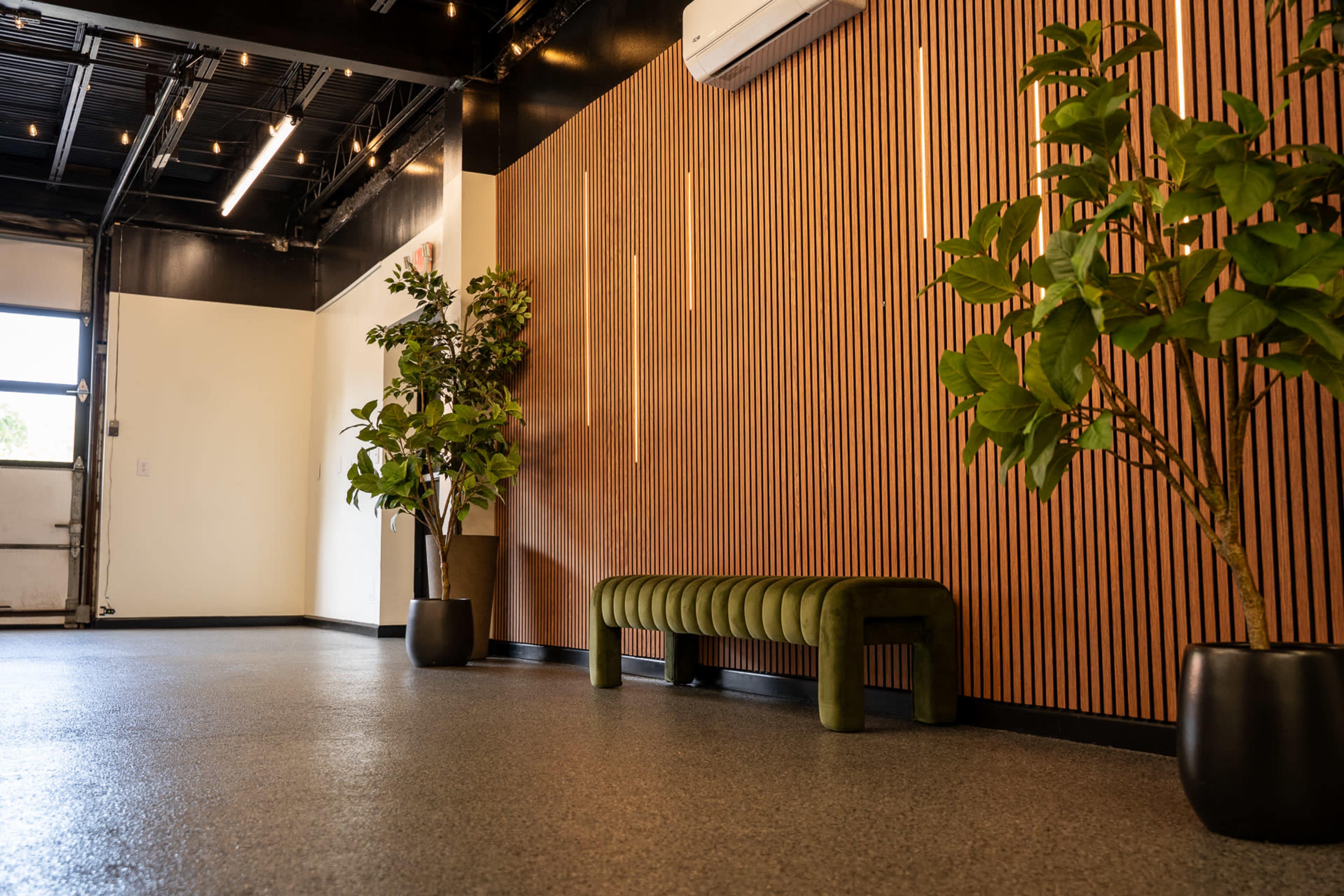 The image shows a modern interior space featuring a wooden slatted wall, two potted plants, and a green upholstered bench.