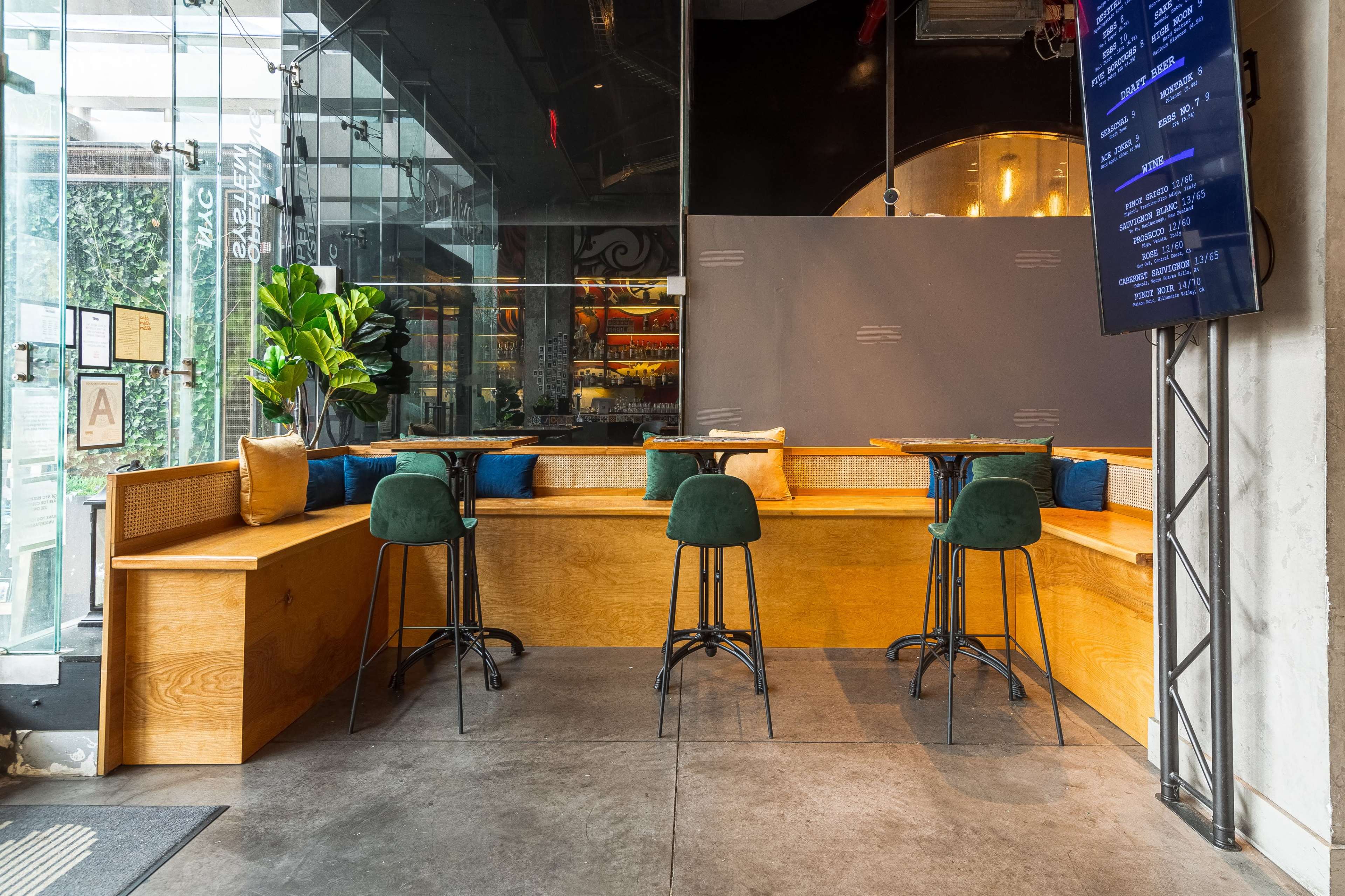 A modern cafe interior featuring a wooden seating area with green barstools, decorative cushions, and large glass windows.