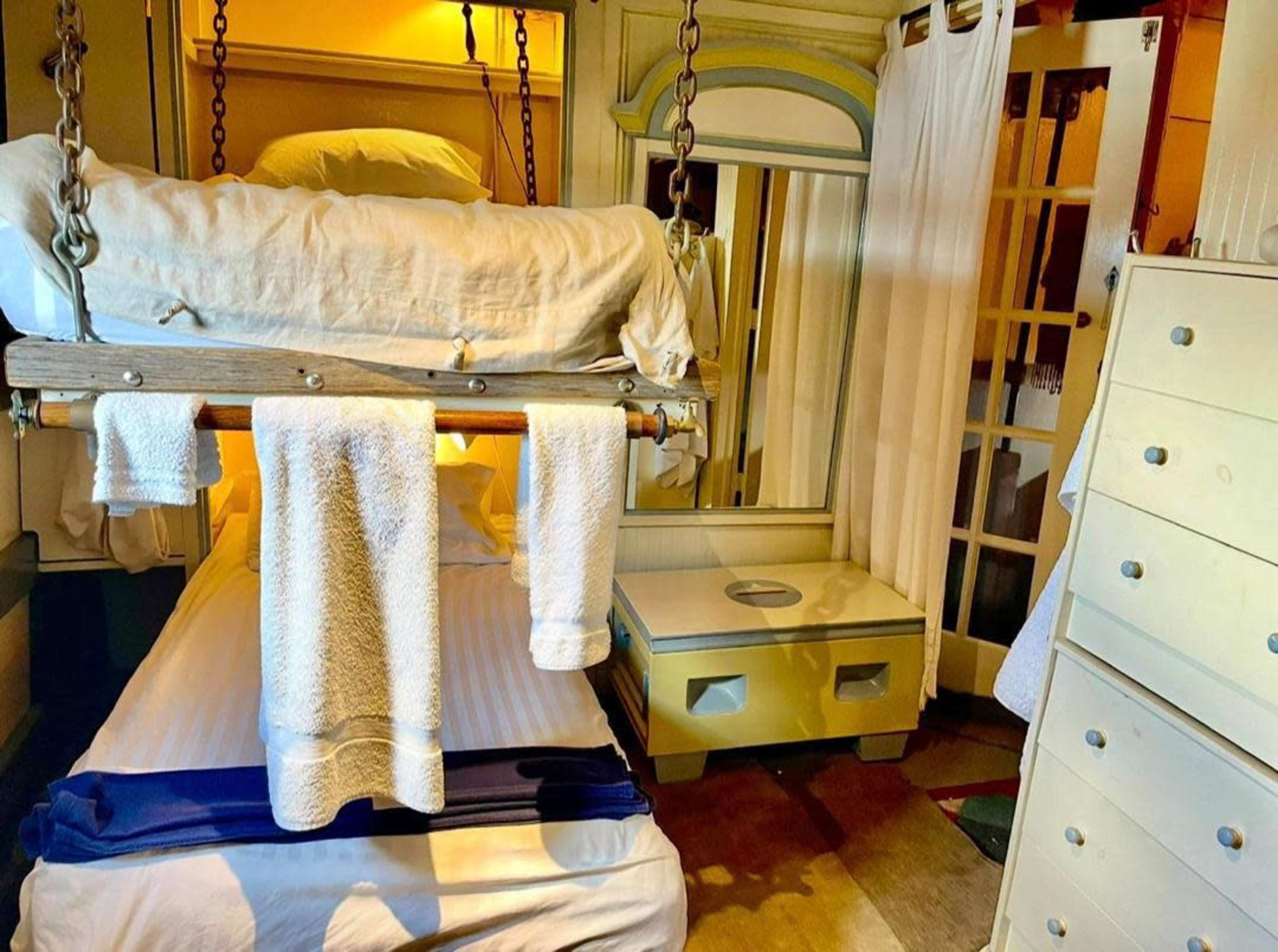 The image shows a small bedroom featuring a suspended bed on chains, with a white sheet and towels hanging nearby, alongside a mirrored wardrobe and a chest of drawers.
