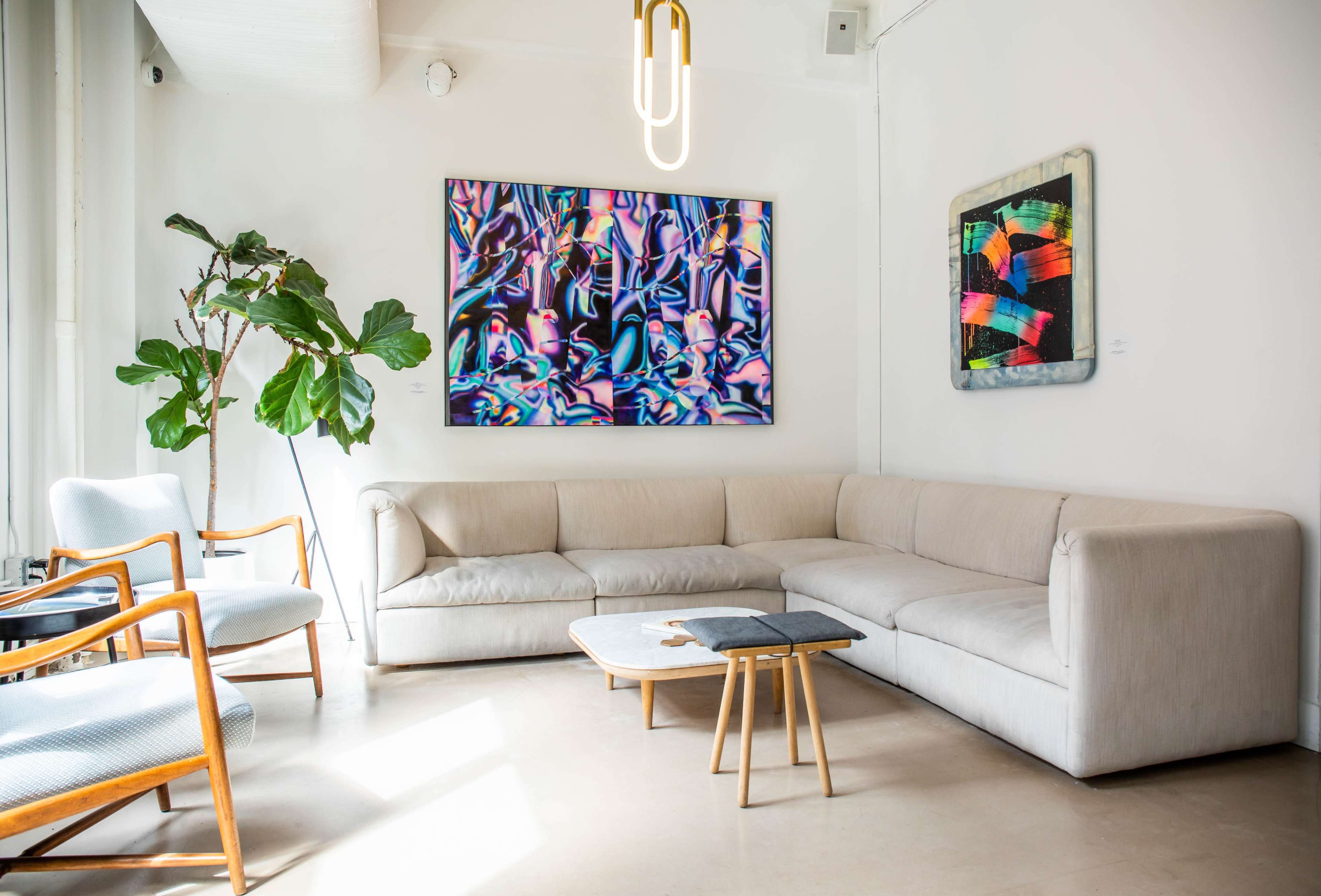 A modern living space featuring a light-colored sectional sofa, two wooden chairs, a small coffee table, and vibrant abstract artwork on the walls.